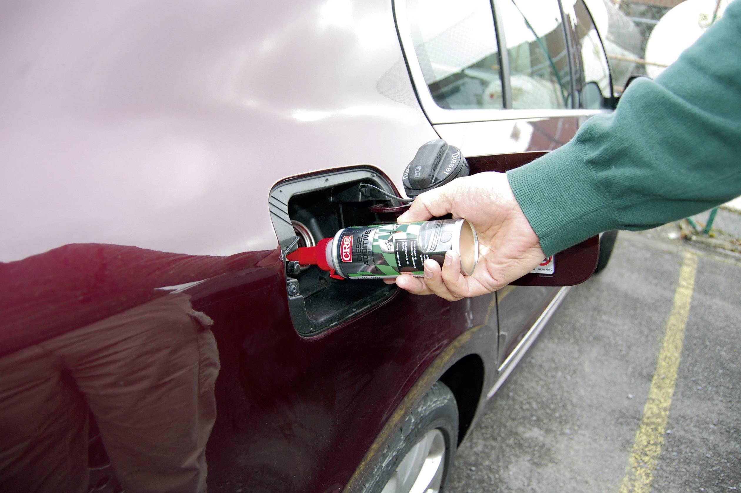 Eine Person füllt ein Additiv aus einer roten Dose in den Kraftstofftank eines weinroten Autos ein.
