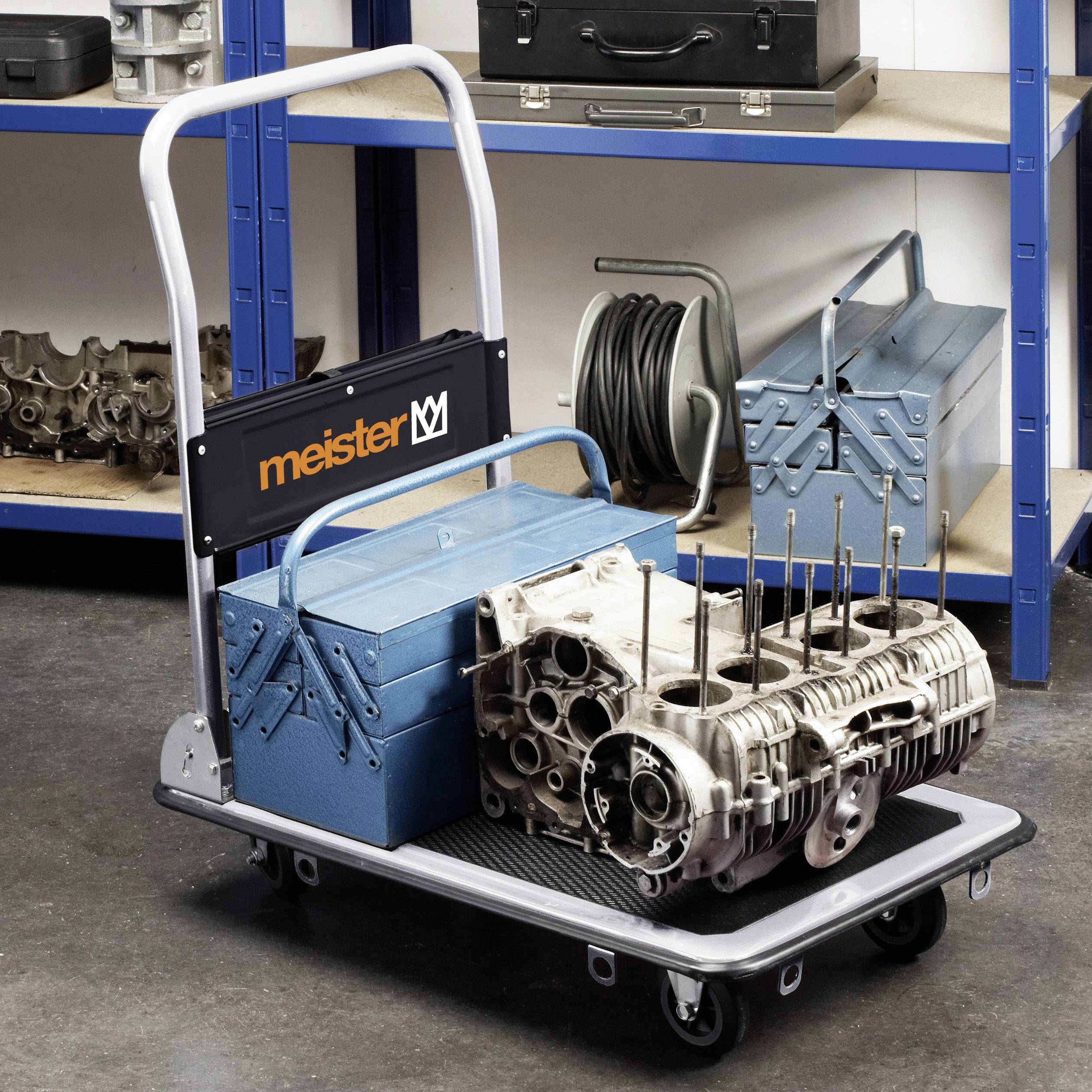 Transportwagen mit Werkzeugkasten und Motorteil, umgeben von Regalen mit Ersatzteilen und Werkzeugen in einer Werkstattumgebung.