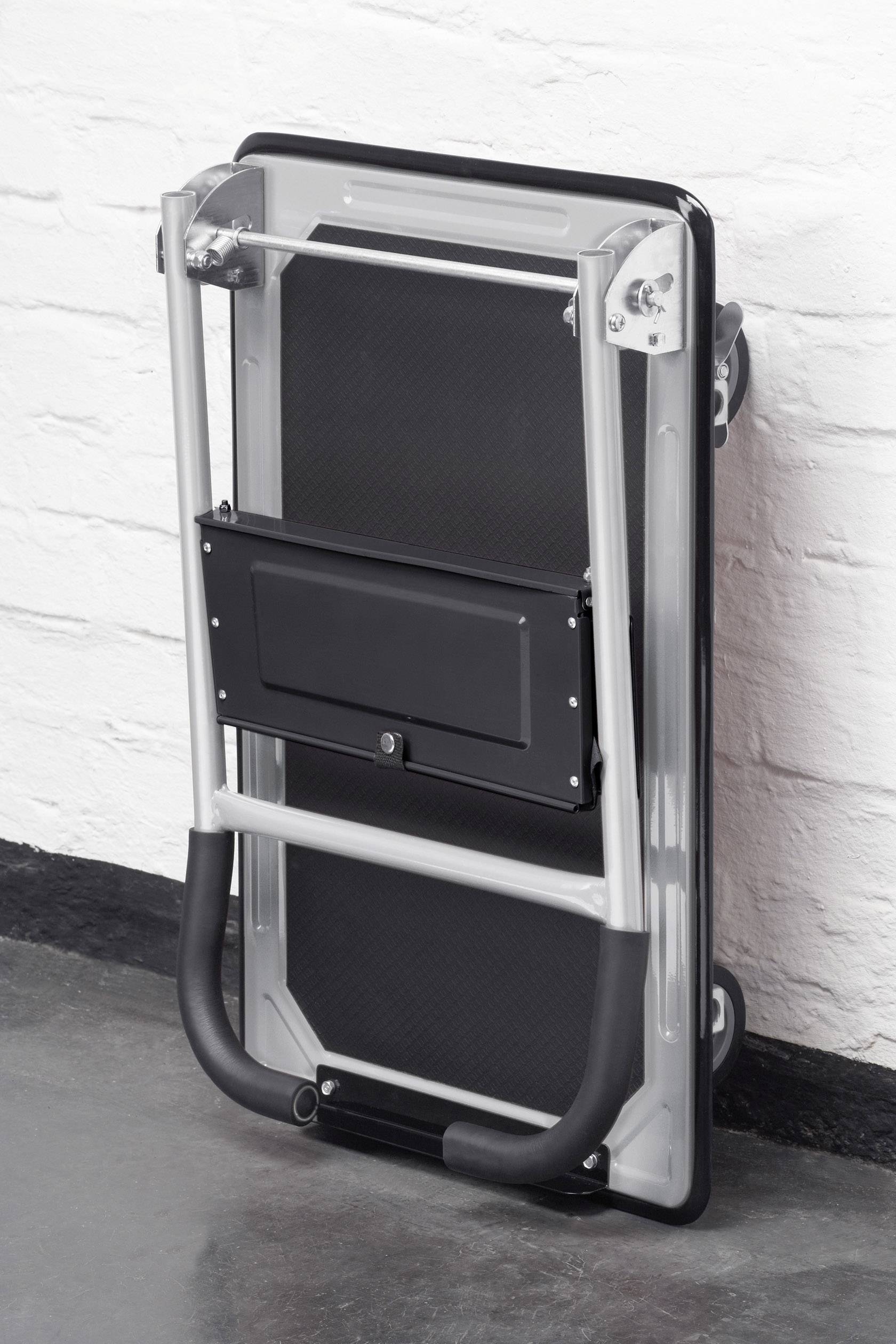 Ein umgeklappter Handwagen lehnt an einer weißen Ziegelwand. Der schwarze und silberne Wagen ist zusammengeklappt und platzsparend verstaut.