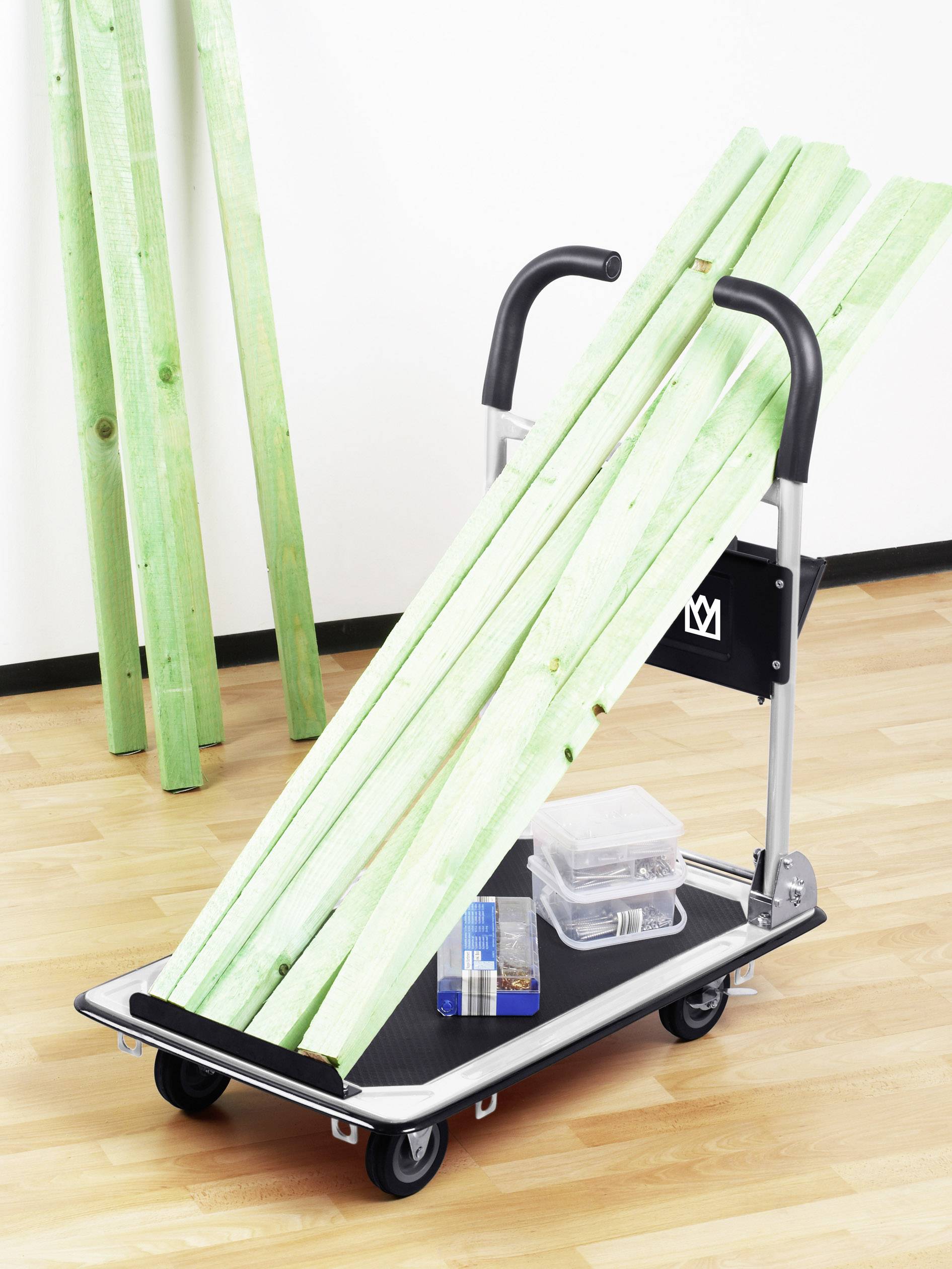 Ein Handwagen aus Metall auf einem Holzboden trägt grüne Holzlatten und eine Plastikbox mit Werkzeug. Im Hintergrund stehen gestapelte Latten an der Wand.
