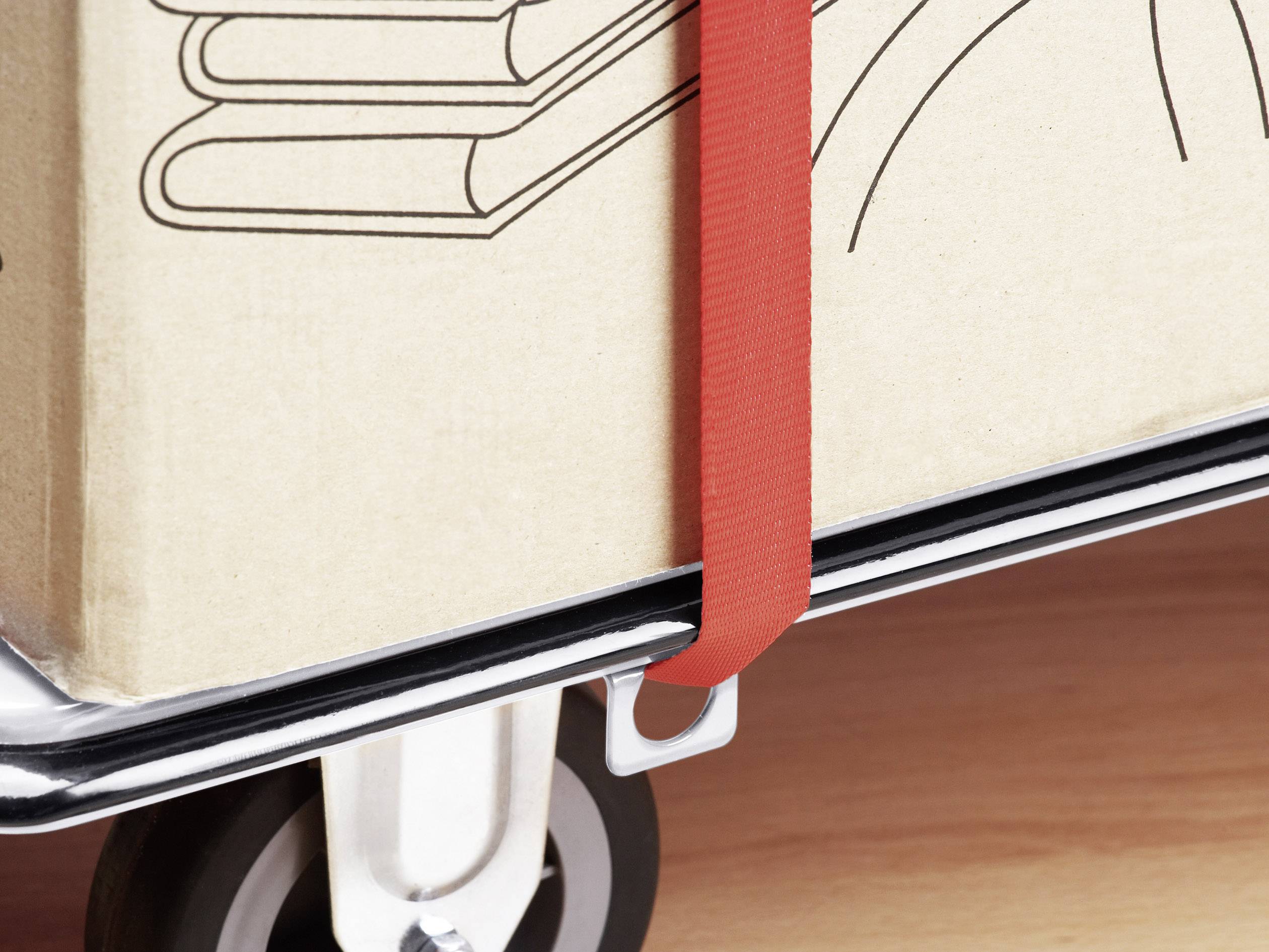 Ein Karton mit Buchsymbolen wird mit einem roten Gurt auf einem Transportwagen gesichert. Der Boden ist aus Holz.