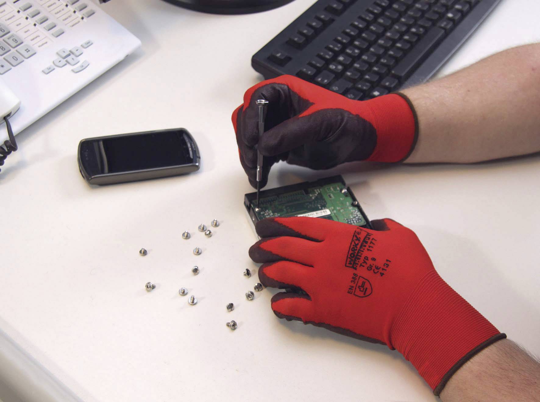 Eine Person repariert eine Computerfestplatte mit Schraubenzieher und Handschuhen auf einem Tisch. Schrauben und ein Smartphone liegen daneben.