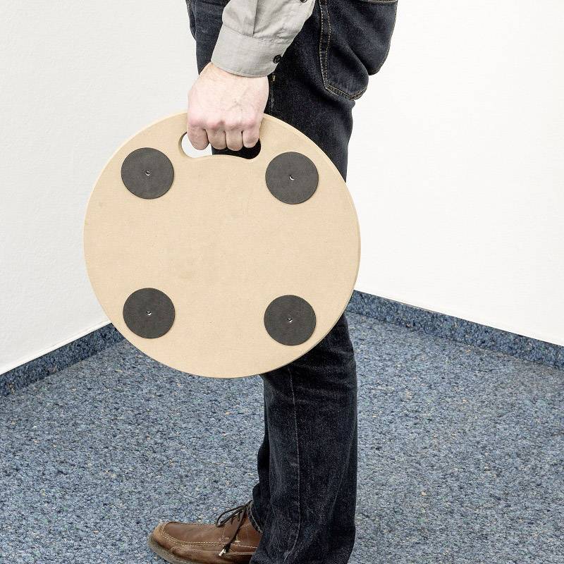 Eine Person hält einen runden Holzdrehteller mit vier schwarzen Rollen, stehend auf einem blau-gemusterten Teppichboden in einem Raum.