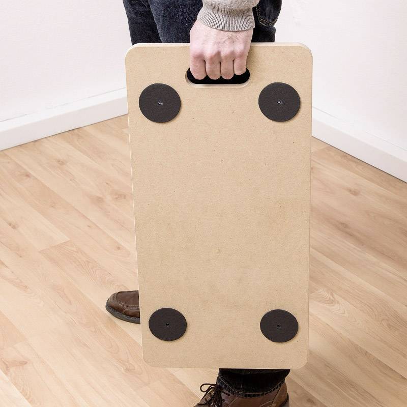 Eine Person trägt ein rechteckiges Balance-Board aus Holz mit schwarzen Anti-Rutsch-Pads auf einer hellen Holzbodenfläche.