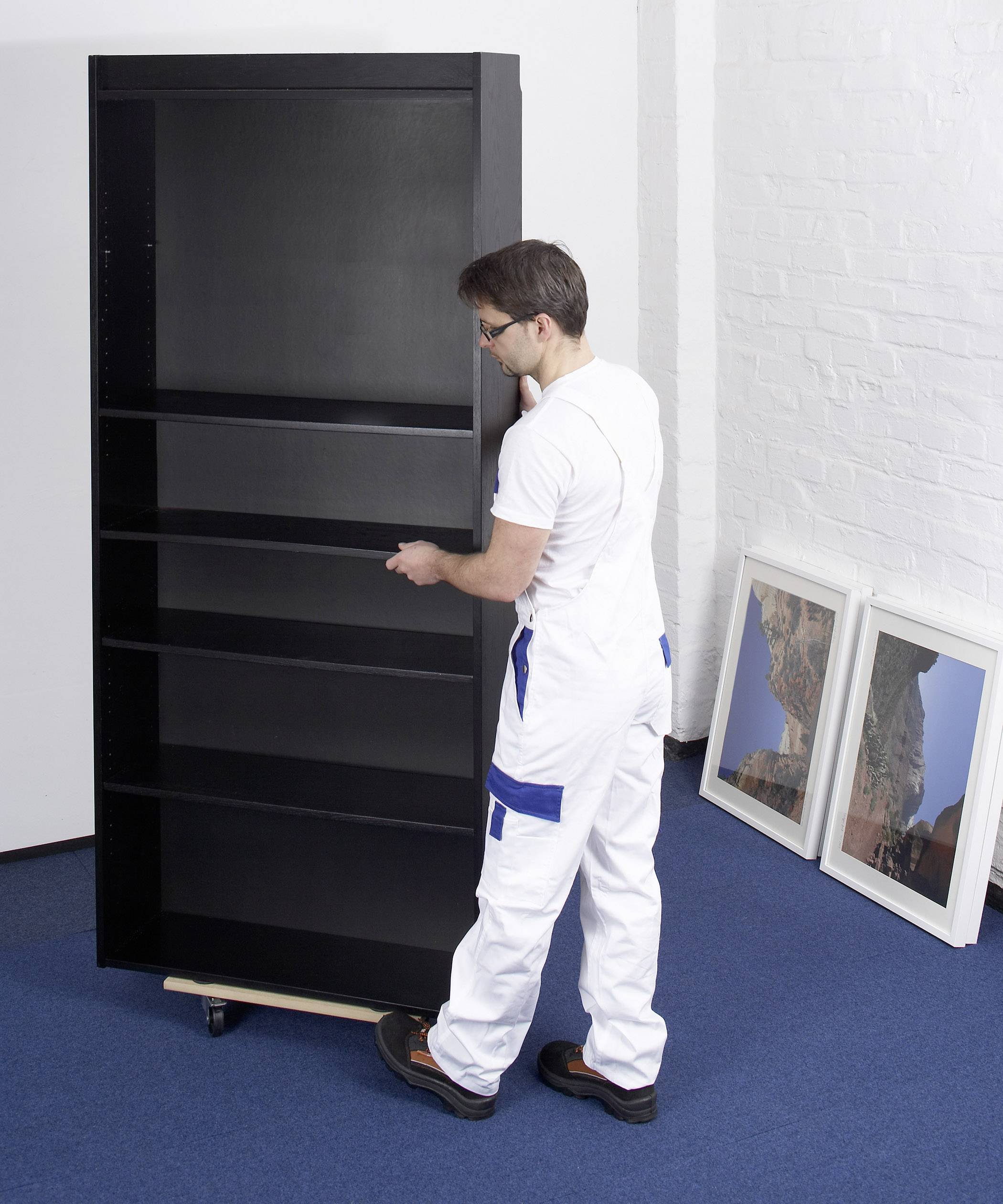 Ein Mann in Arbeitskleidung baut ein großes, schwarzes Regal auf einem blauen Teppich neben zwei gemalten Bildern an der Wand zusammen.