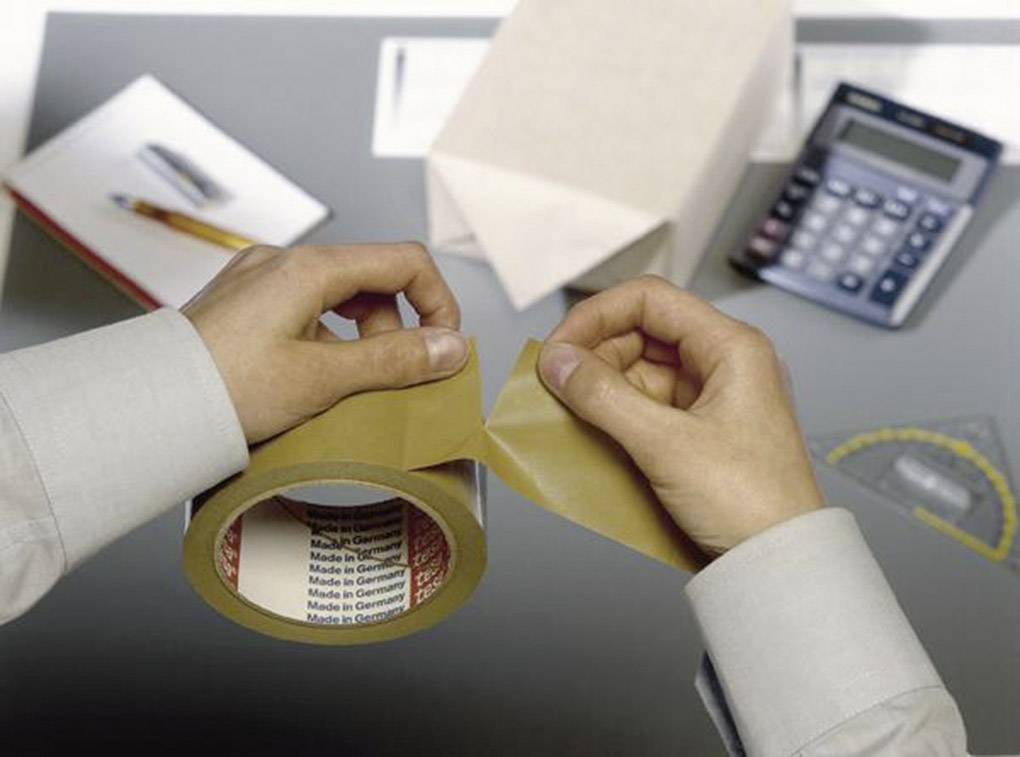 Eine Person zieht Klebeband von einer Rolle auf einem Schreibtisch ab. Neben der Rolle liegen eine Schachtel, ein Taschenrechner und Schreibwaren.