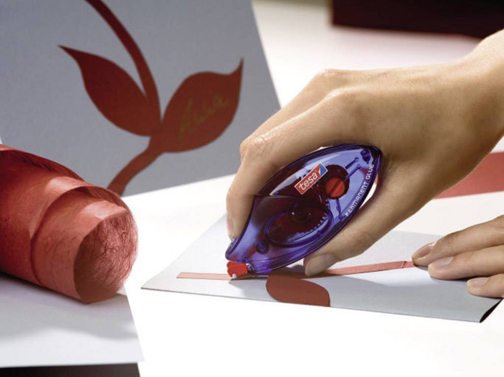 Eine Hand benutzt einen Korrekturroller, um eine rote Linie auf grauem Papier zu markieren. Im Hintergrund befindet sich ein rotes Blattmuster.