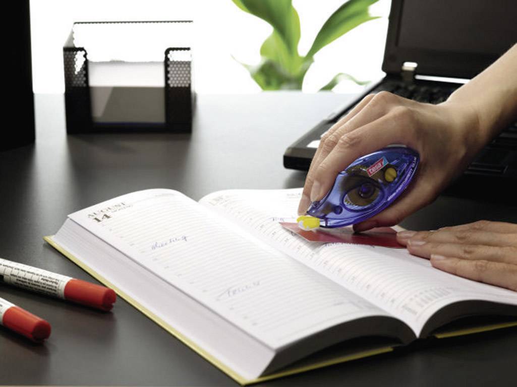 Eine Person korrigiert handgeschriebene Notizen in einem Terminkalender mit einem Korrekturroller auf einem Schreibtisch.