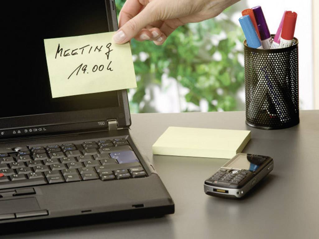 Eine Hand legt einen Notizblock mit der Aufschrift 'Meeting 19:00' auf einen Laptop. Daneben liegen ein Handy und bunte Stifte.