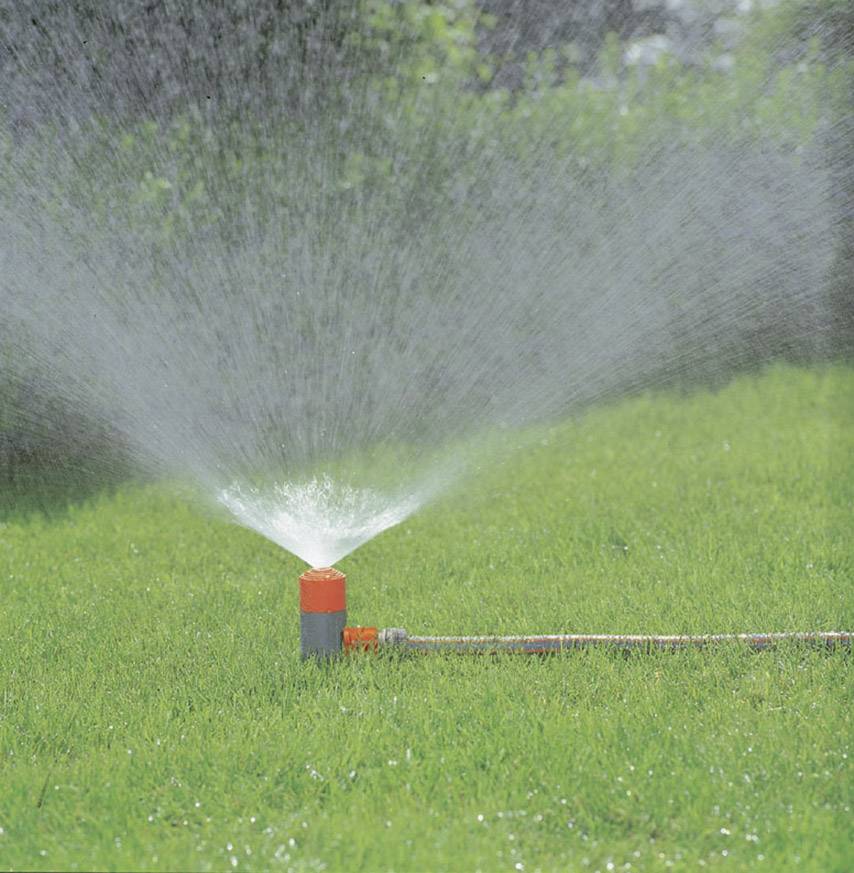Rasenfläche wird von einem Sprinkler bewässert. Wasserstrahlen verteilen sich gleichmäßig über das grüne Gras im Garten.