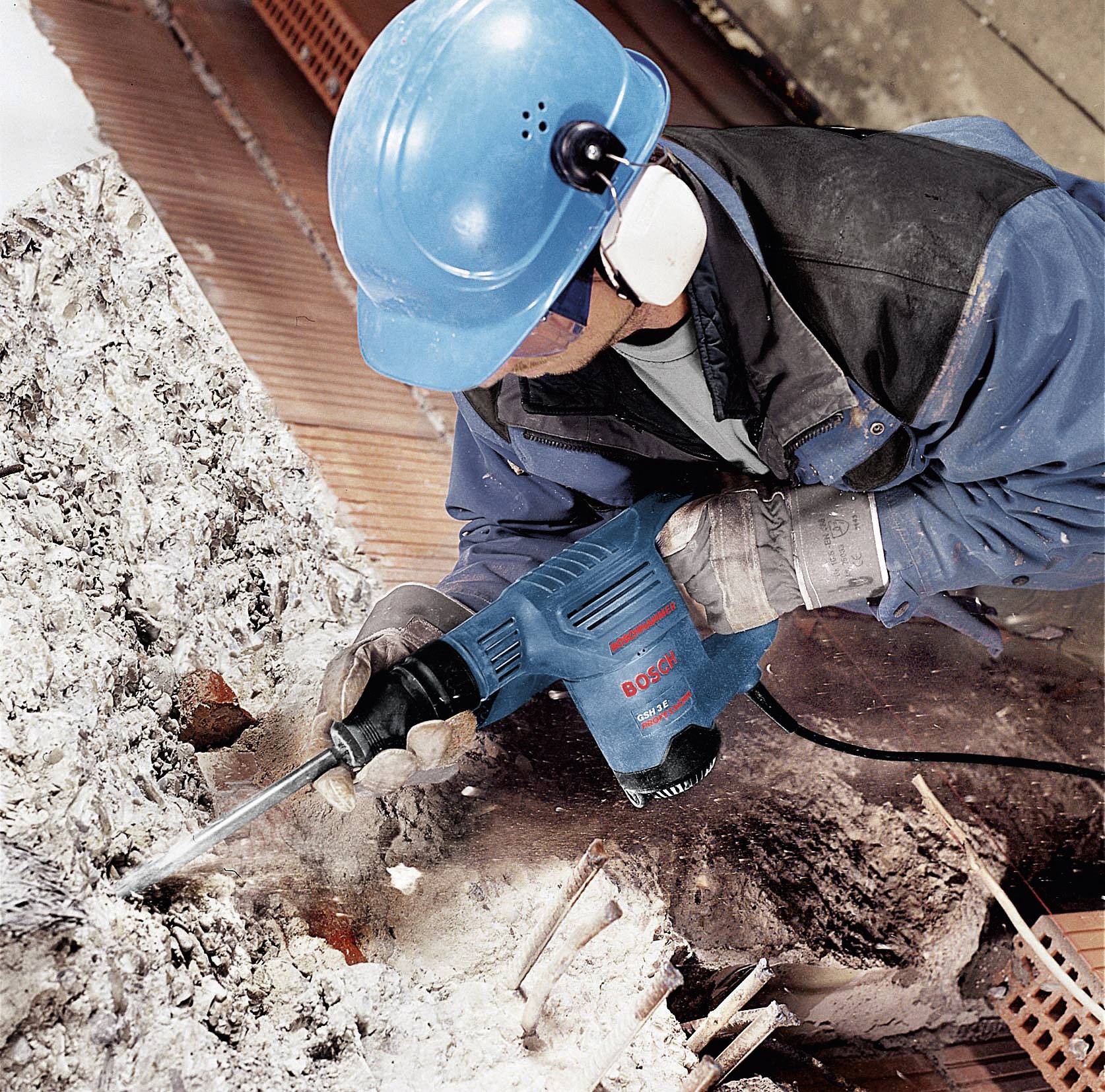 Eine Person mit Schutzhelm und Gehörschutz bohrt mit einer blauen Bohrmaschine in Betontrümmer auf einer Baustelle.