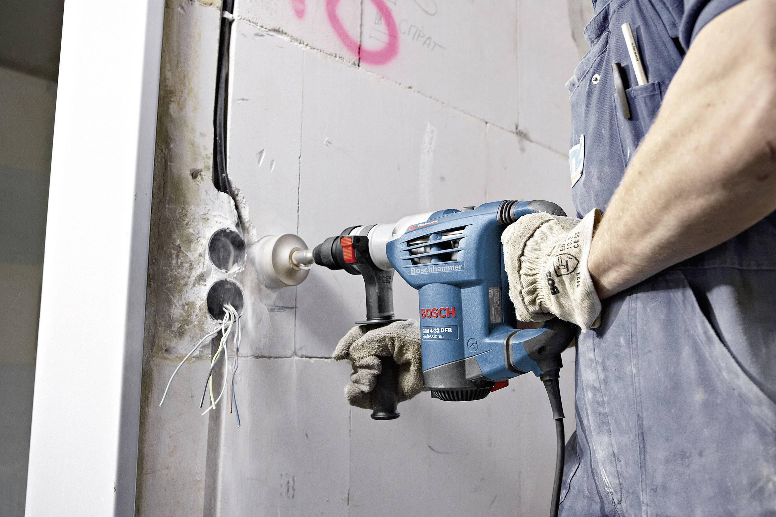 Eine Person in Arbeitskleidung bohrt mit einer Bohrmaschine ein Loch in eine Wand, aus der Kabel herausragen.