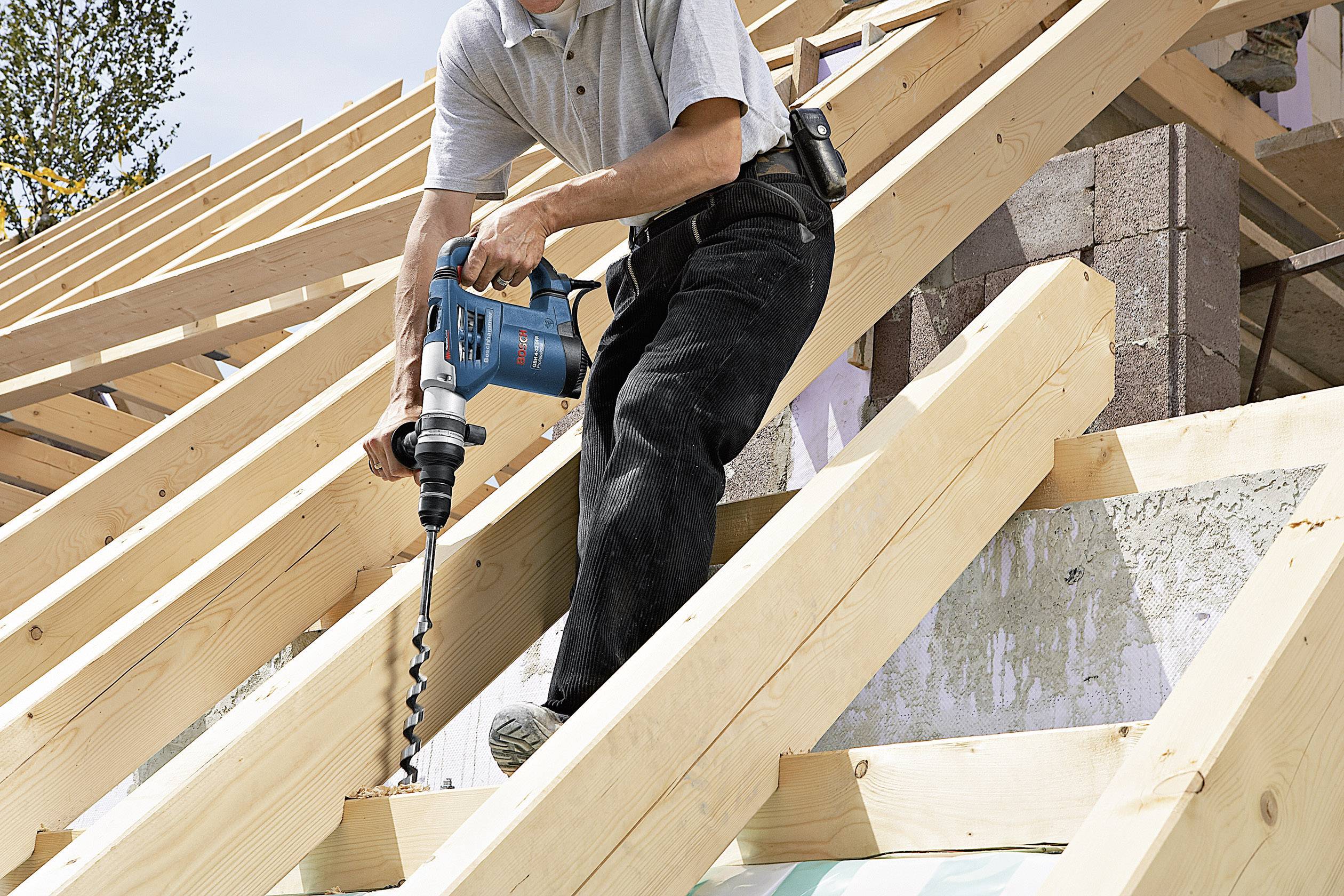 Ein Bauarbeiter bohrt mit einem blauen Werkzeug ein Loch in die Holzbalken eines Dachgerüsts. Im Hintergrund sind Ziegelsteine zu sehen.