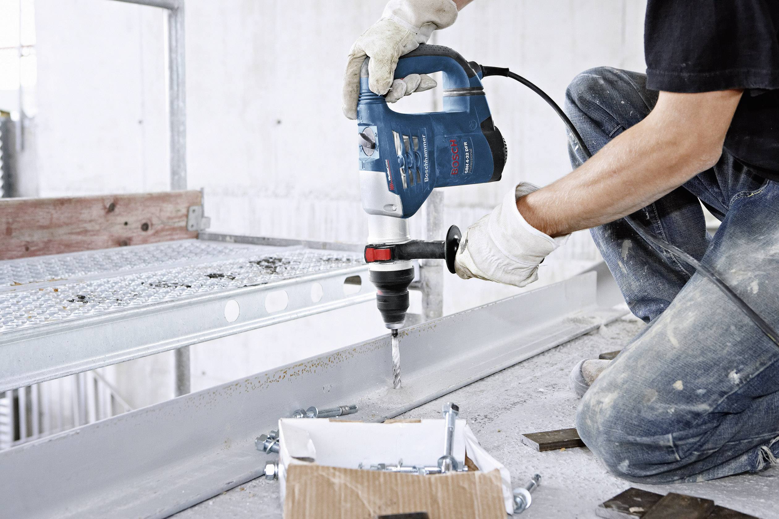 Eine Person bohrt mit einer blauen Bohrmaschine in einen Metallträger. Auf dem Boden liegt eine Schachtel mit Schrauben und Werkzeugen.