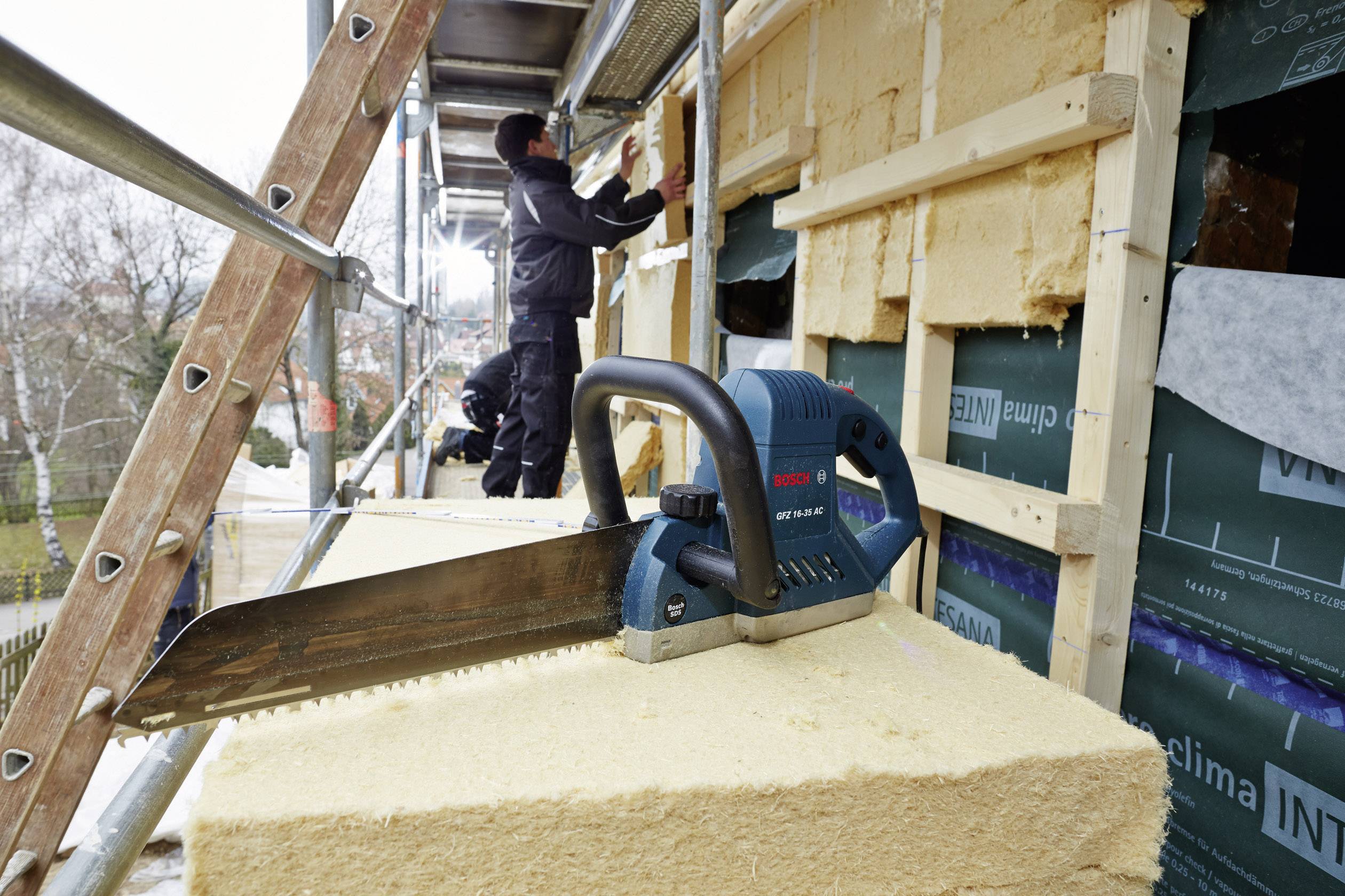 Ein Arbeiter isoliert die Fassade eines Gebäudes mit Dämmmaterial, während im Vordergrund eine elektrische Säge liegt.