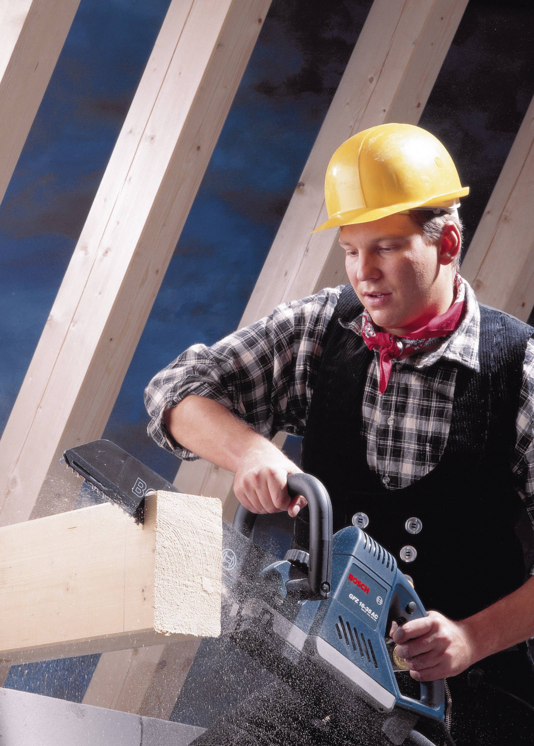 Ein Bauarbeiter in Schutzkleidung sägt mit einer Kettensäge ein Holzbrett. Hintergrund zeigt Holzstreben und blauen Himmel.