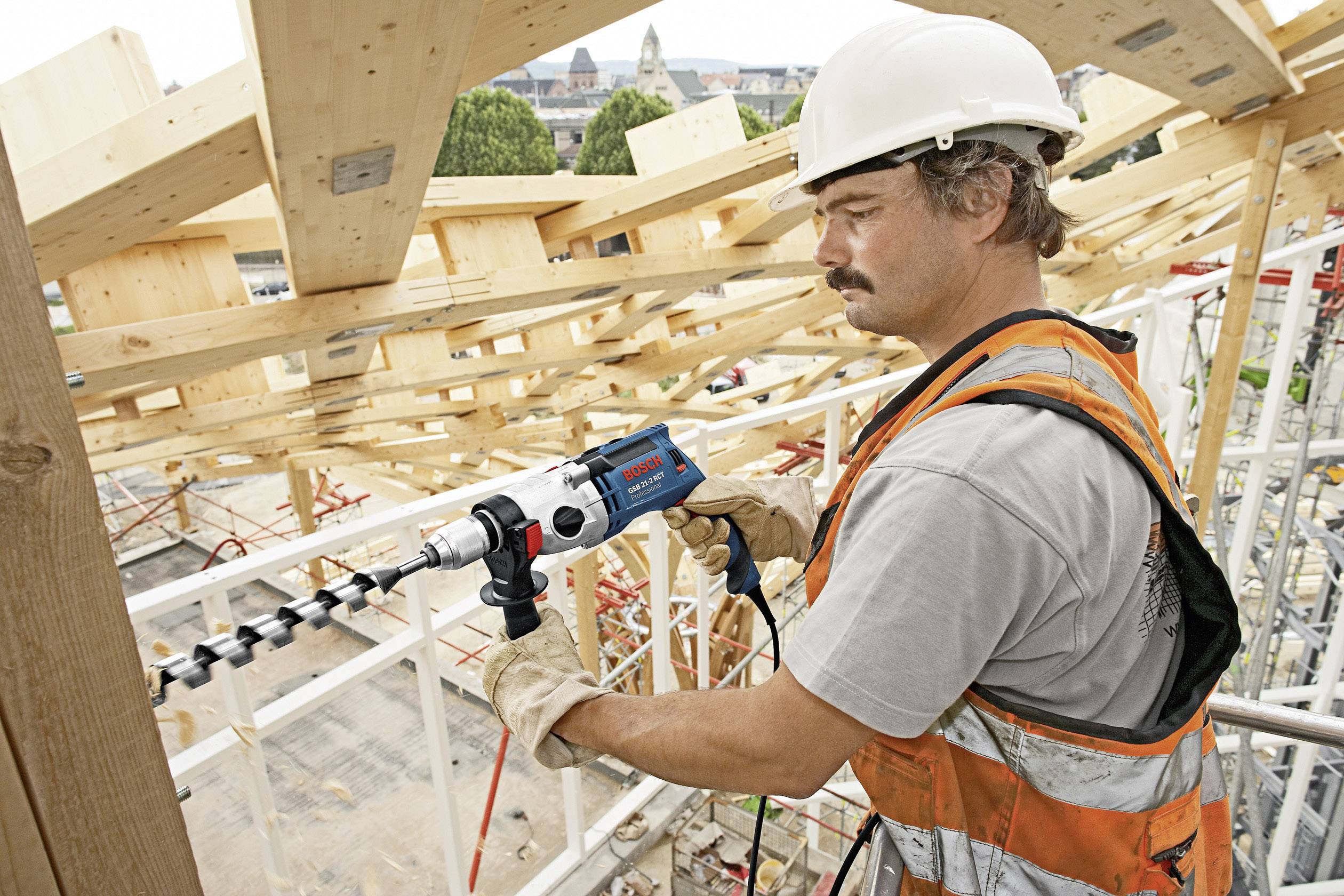 Ein Bauarbeiter in Schutzhelm und Weste arbeitet auf einer Holzbaustelle mit einem Bohrer. Im Hintergrund sind Gebäude zu sehen.