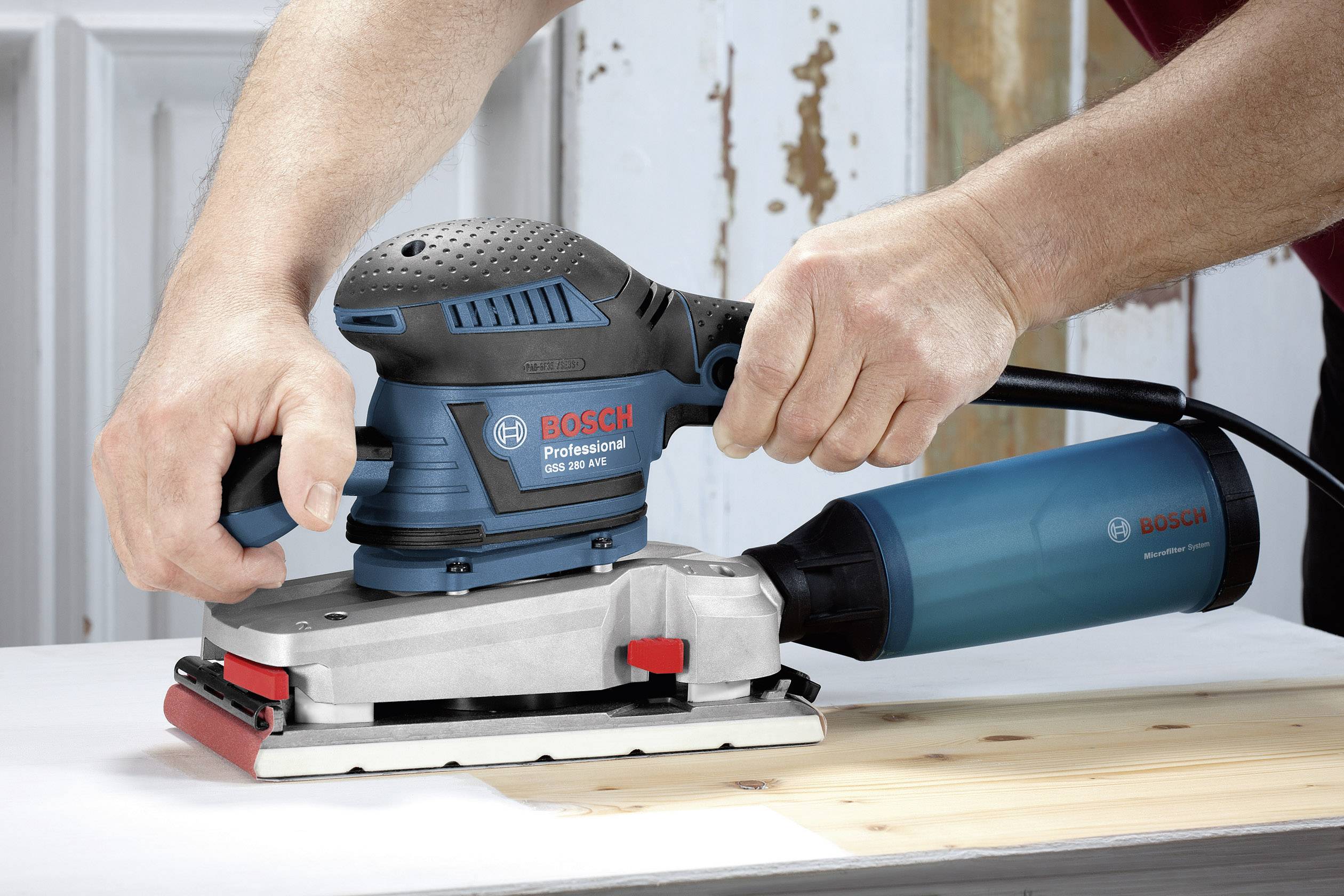 Eine Person benutzt einen Bosch Professional Schwingschleifer auf einer Holzoberfläche. Im Hintergrund sind verwitterte Holzpaneele zu sehen.