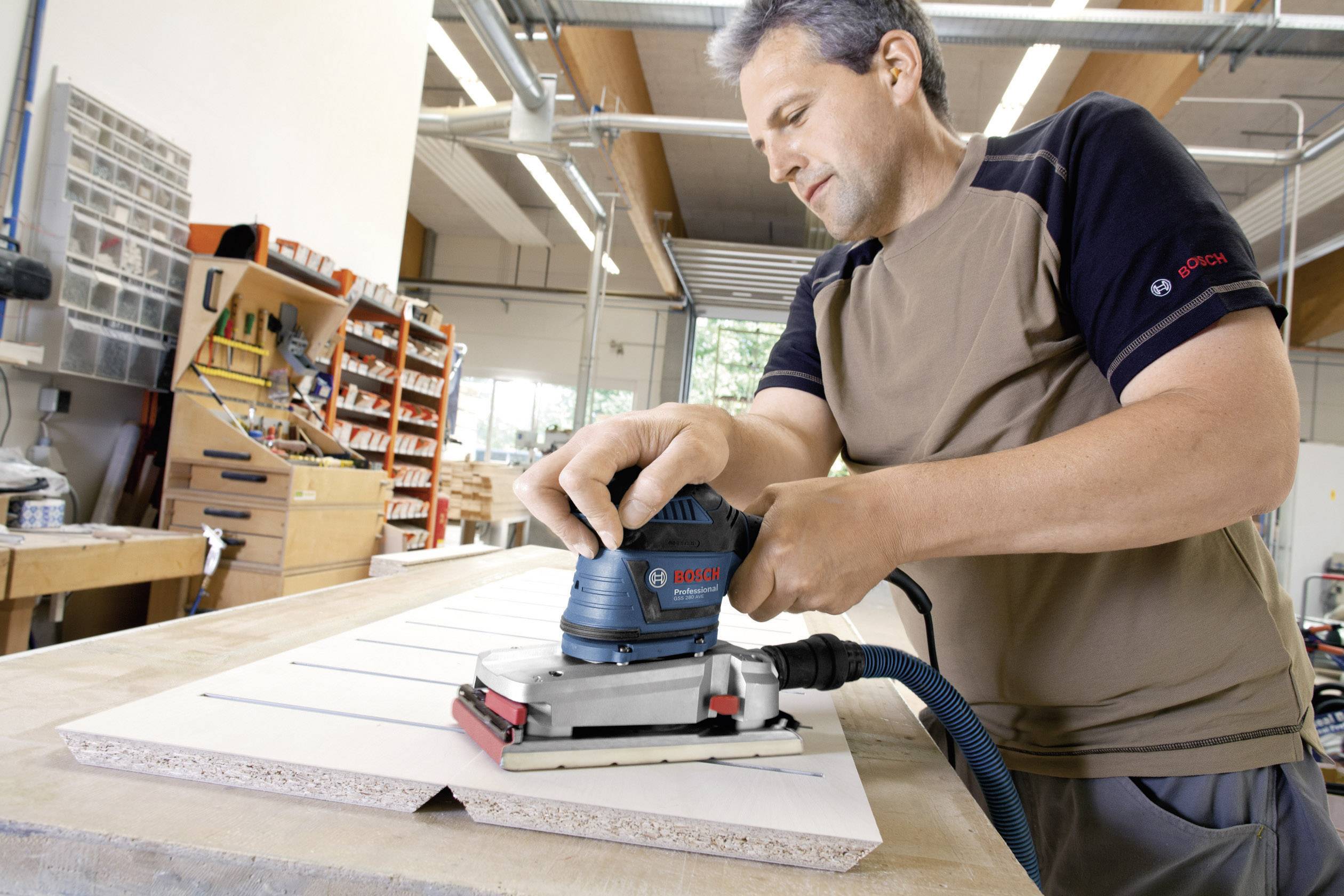 Ein Mann benutzt ein elektrisches Schleifgerät, um eine Holzplatte in einer Werkstatt zu bearbeiten. Im Hintergrund sind Werkzeuge sichtbar.