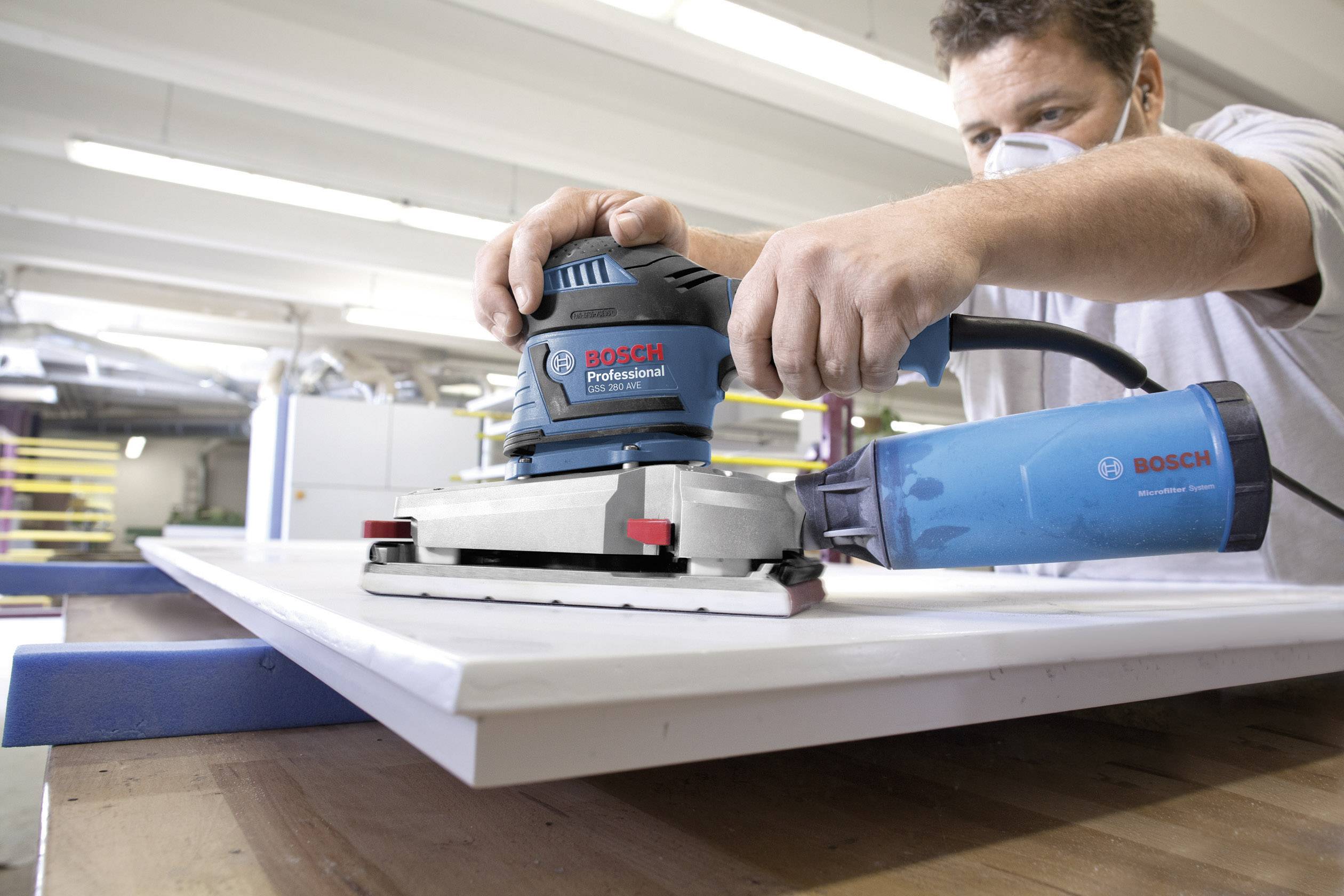 Ein Mann mit Atemmaske benutzt einen blauen Bosch Professional Schwingschleifer, um eine weiße Holzplatte in einer Werkstatt zu bearbeiten.