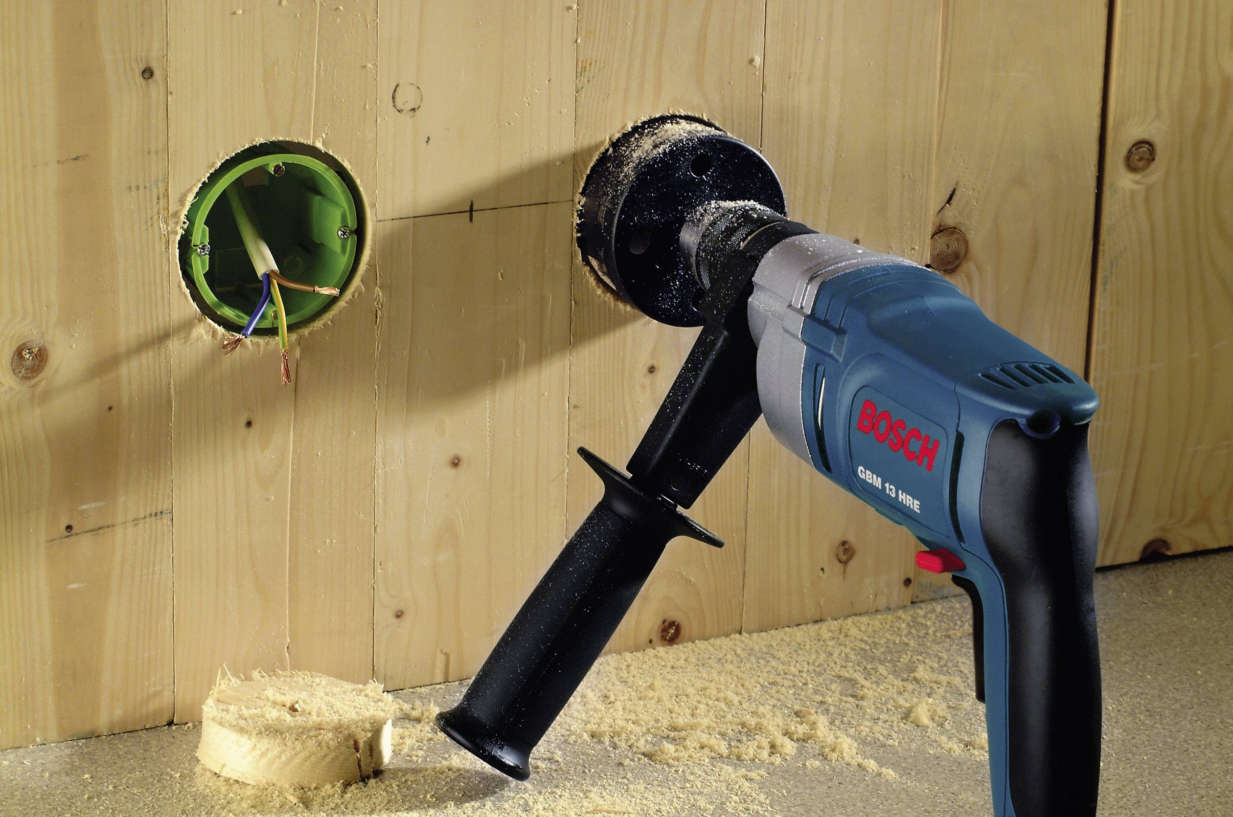 Bohrmaschine mit Lochsäge am Bohrer erzeugt rundes Loch in Holzplatte neben elektrischer Anschlussdose.