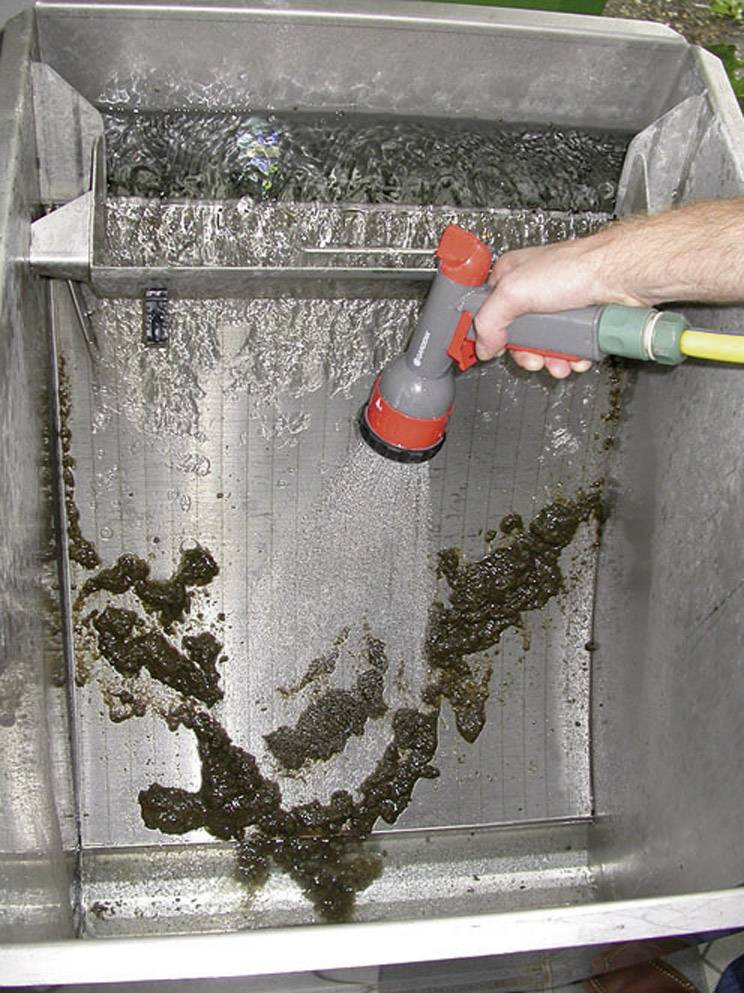 Person spritzt Schmutz mit einem Gartenschlauch von einer Metalloberfläche ab, das Wasser fließt in einen Ablass im Becken.