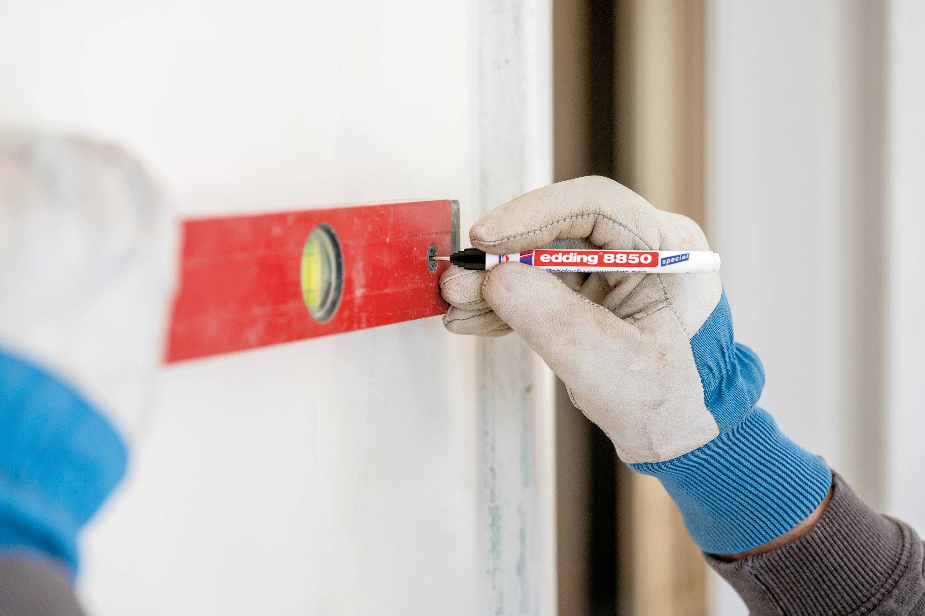 Eine Person zeichnet mit einem roten Marker auf einer Wand, nutzt eine rote Wasserwaage zum Ausrichten, trägt Handschuhe.