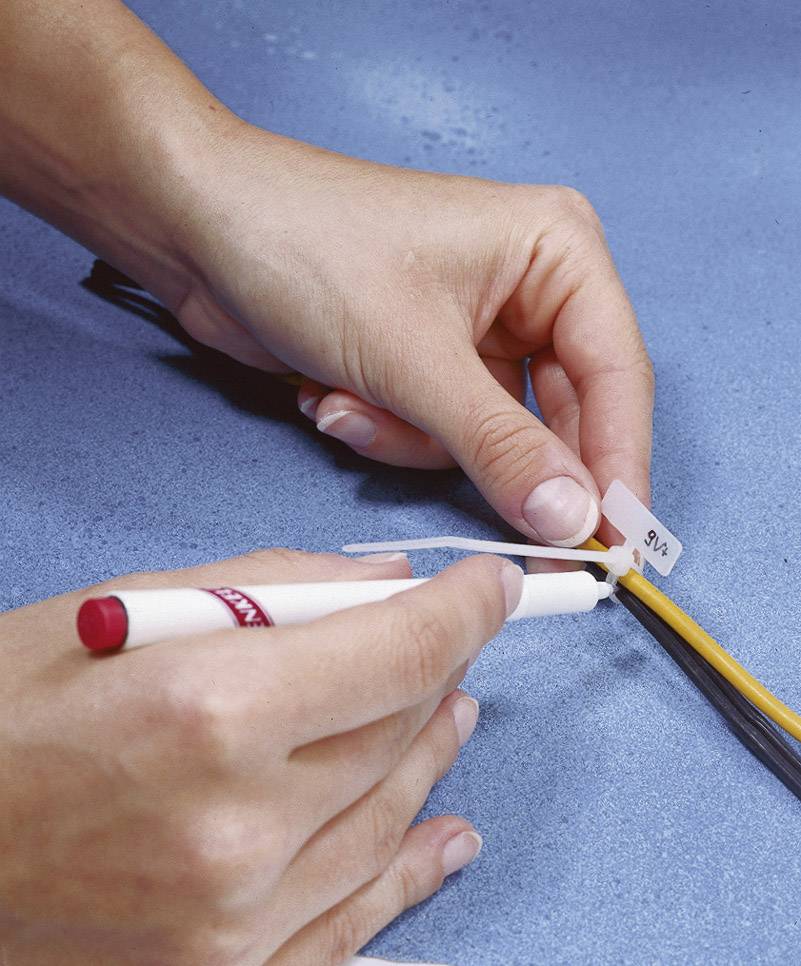 Zwei Hände beschriften ein Kabel mit einem weißen Etikett und einem roten Stift auf einer blauen Oberfläche.