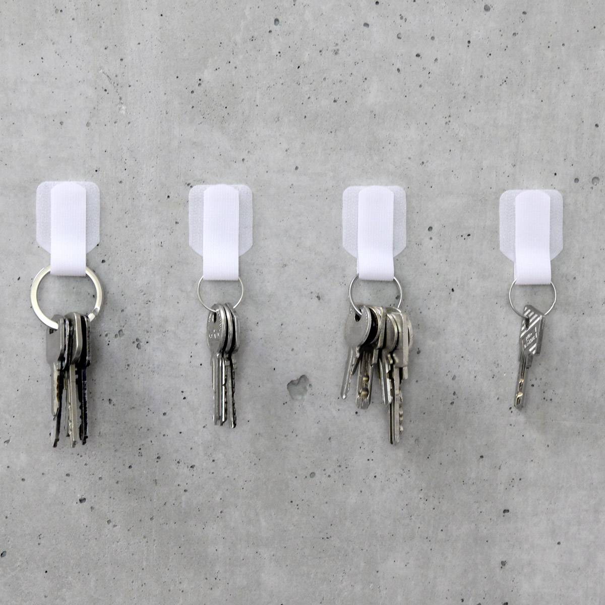 Vier weiße Haken an der Wand, jeweils mit einem Bund Schlüssel daran hängend. Die Haken sind an einer Betonwand befestigt.