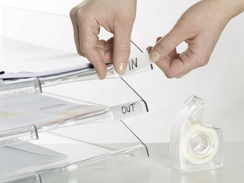 Eine Person legt Dokumente in ein dreiteiliges Ablagesystem mit den Etiketten 'IN' und 'OUT'. Ein Klebebandabroller ist daneben.