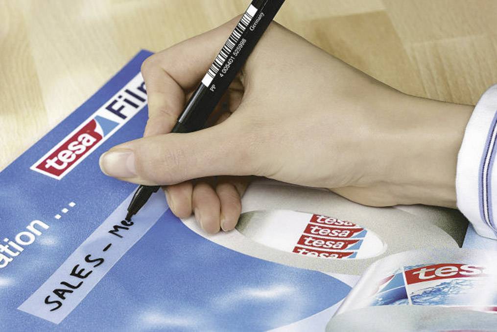 Eine Hand schreibt mit einem schwarzen Stift auf Papier mit 'tesa'-Logo und Aufschrift 'SALES - MEETING'.