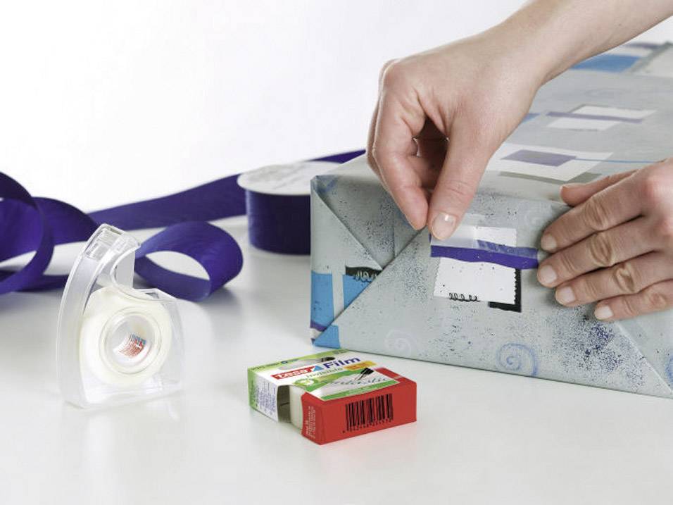 Eine Person verpackt ein Geschenk mit blauem Geschenkband und Klebeband. Auf dem Tisch sind Klebeband und eine Filmpackung sichtbar.