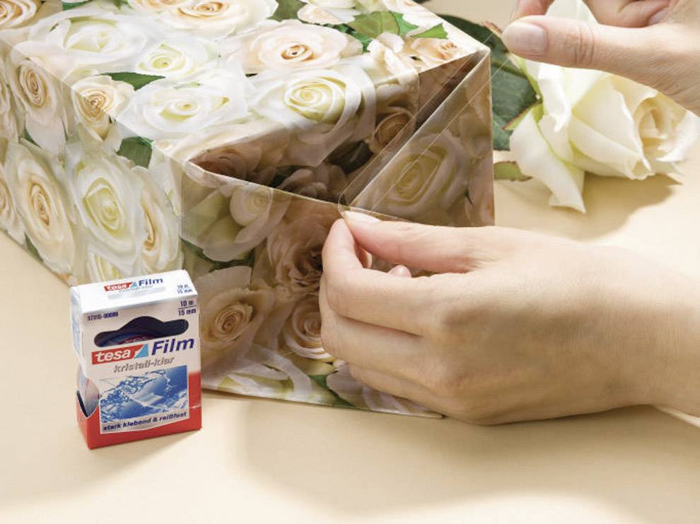 Eine Person verpackt ein Geschenk mit Rosenpapier und nutzt Klebeband. Neben der Hand liegt eine Packung Tesa-Film.