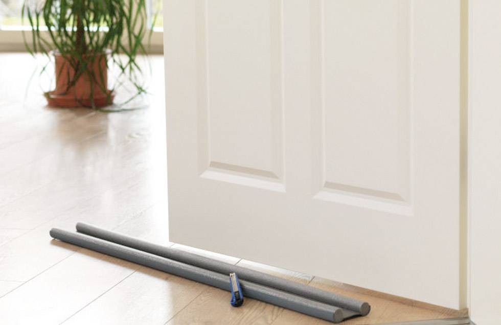 Eine teilweise geöffnete weiße Tür, am Boden mit grauen Isolierrohren und einem Teppichmesser. Im Hintergrund steht eine Pflanze.