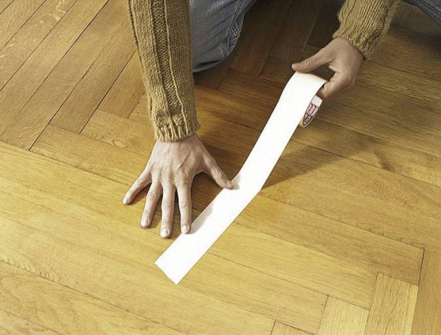 Person legt ein Stück Klebeband auf einen Parkettboden.