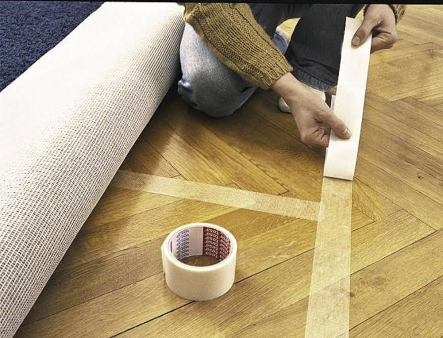 Jemand klebt Teppichklebeband auf einen Holzboden, um einen Teppich zu verlegen. Im Hintergrund liegt ein aufgerollter Teppich.