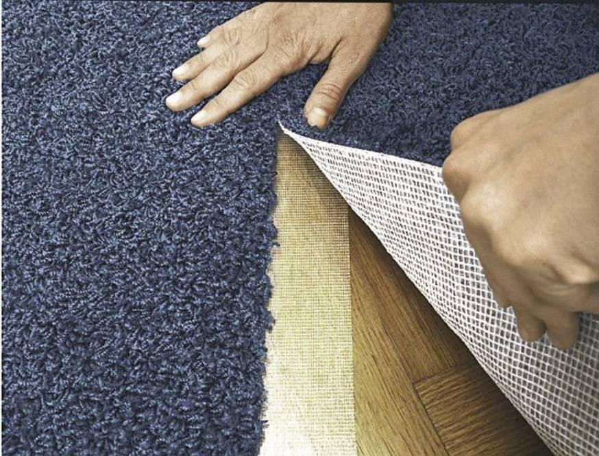 Person legt einen blauen Teppich auf einen Holzboden, indem sie die Kanten glättet und den Teppich sorgfältig ausrollt.