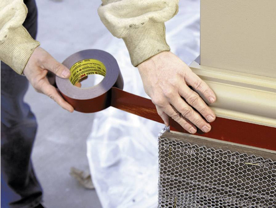 Person klebt ein rotes Band um eine Säule. Hände halten das Band fest. Der Hintergrund zeigt unscharfe Bauutensilien.