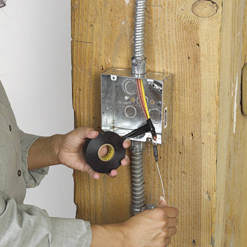 Eine Person installiert ein elektrisches Kabel in eine Metallbox an einer Wand, verwendet schwarzes Klebeband zur Befestigung.