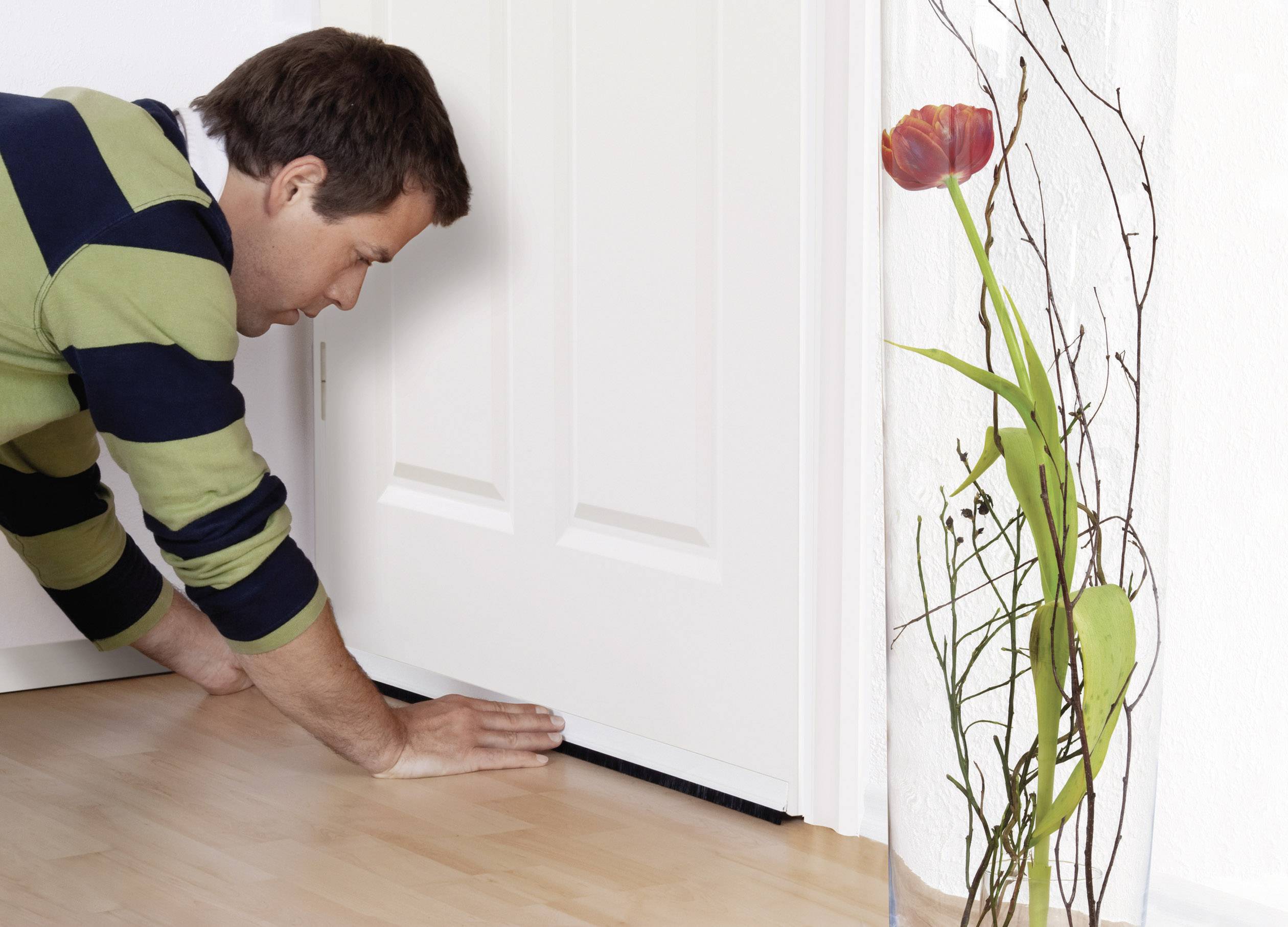 Ein Mann installiert eine Türdichtung an einer weißen Tür, um Zugluft zu verhindern. Daneben steht eine Vase mit einer roten Blume.