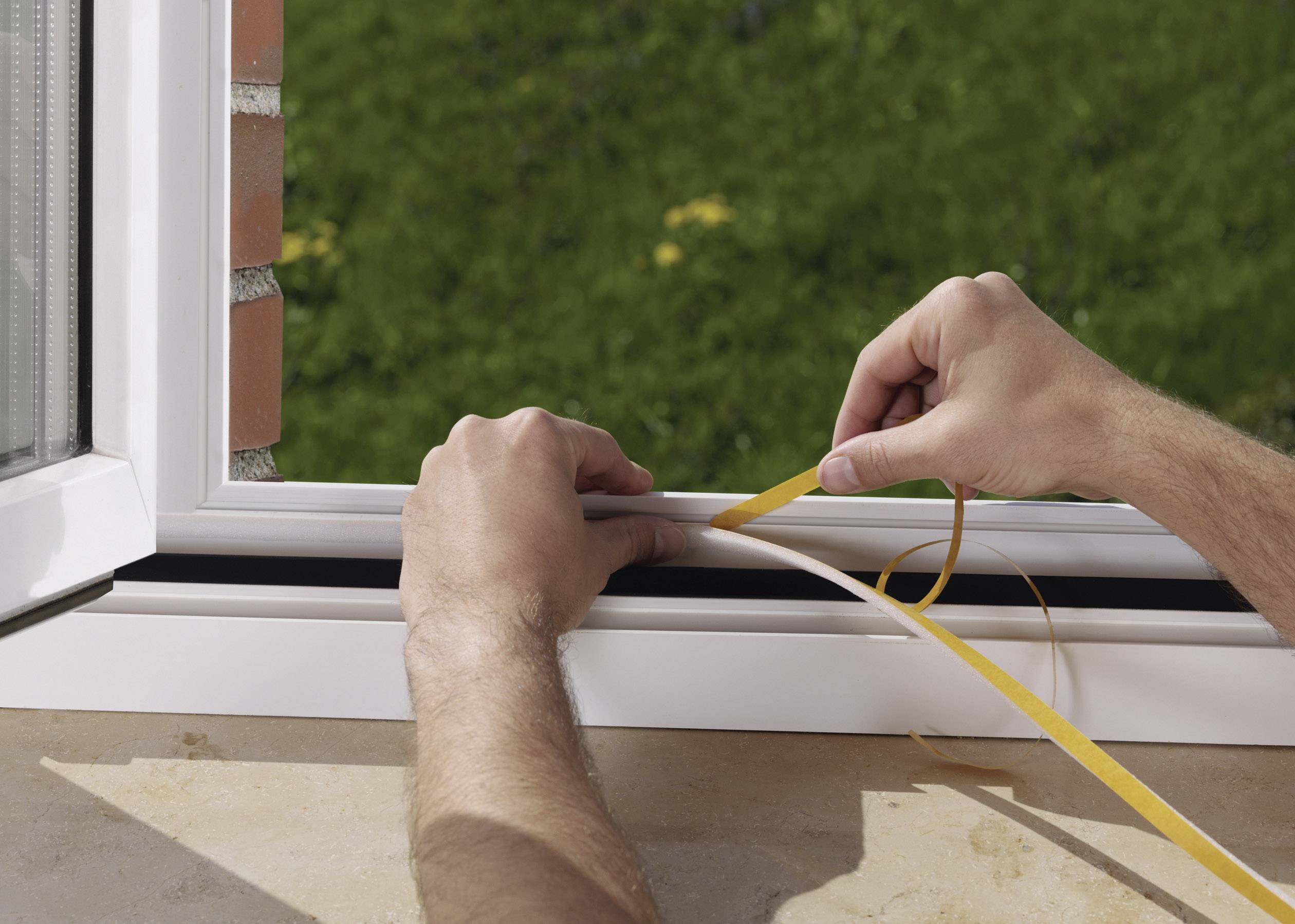 Hände eines Menschen, der ein Dichtungsband an einem offenen Fensterrahmen im Tageslicht anbringt, mit grünem Rasen im Hintergrund.
