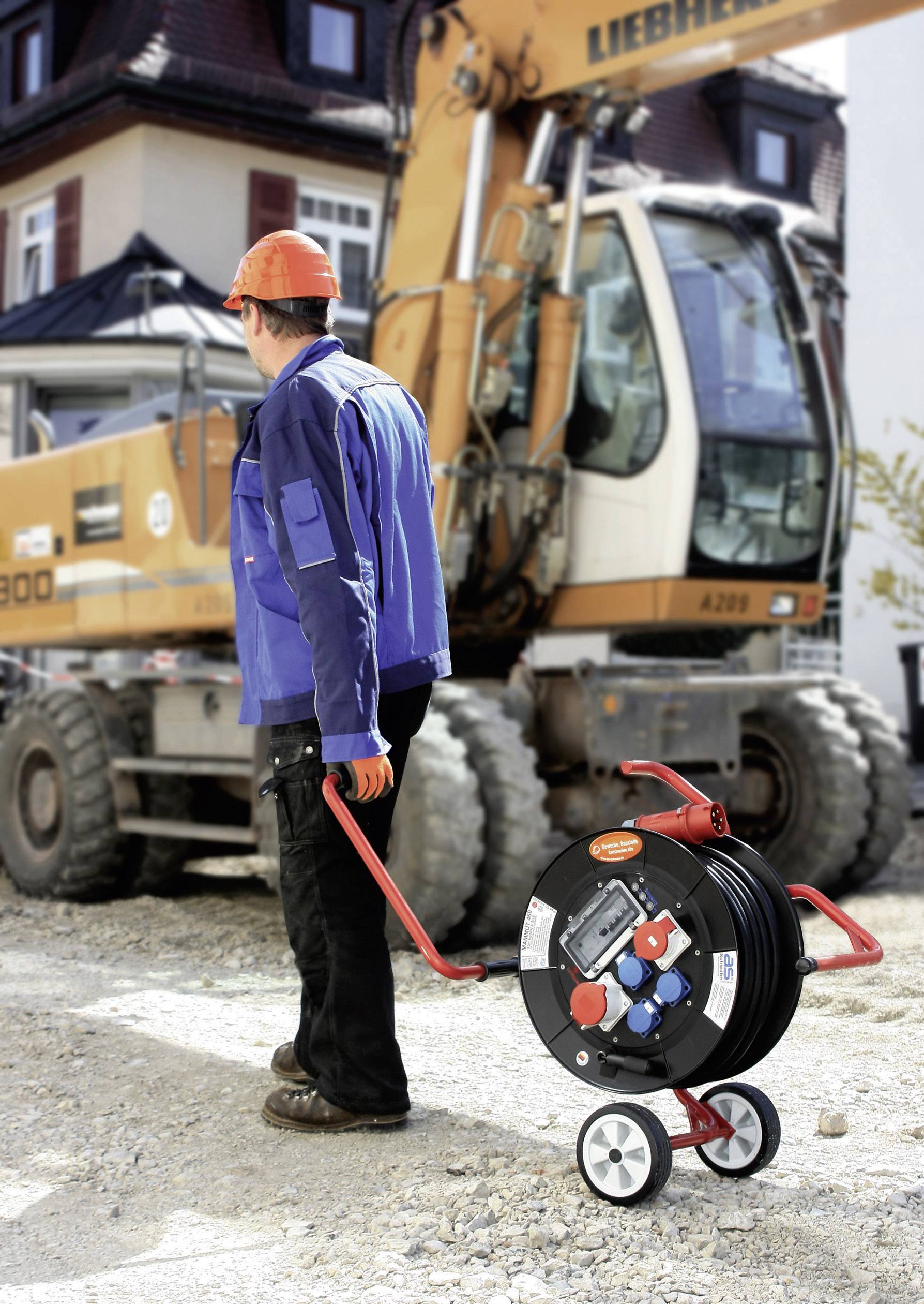 Bauarbeiter mit Helm zieht eine Kabeltrommel auf einer Baustelle, im Hintergrund ein Bagger.