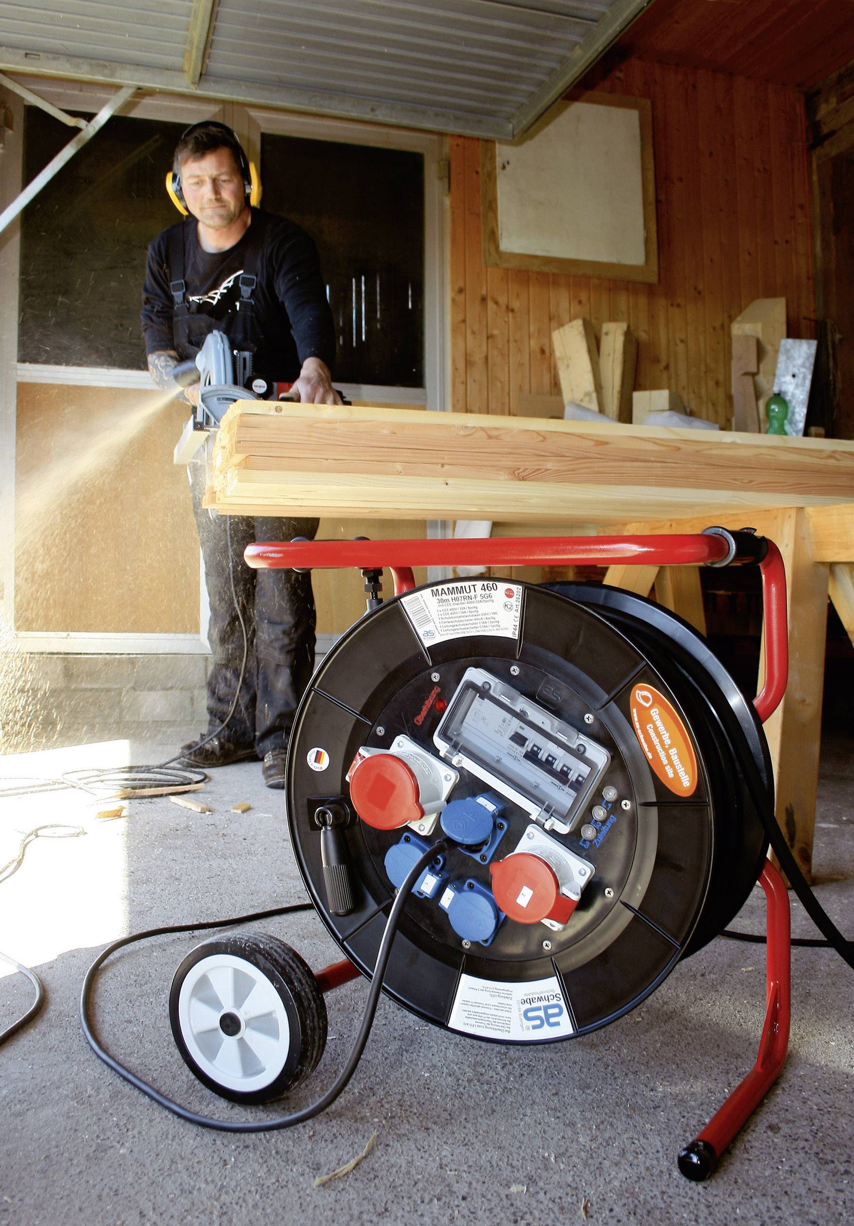 Ein Handwerker mit Gehörschutz sägt Holz an einem Tisch in einer Werkstatt. Im Vordergrund befindet sich eine Kabeltrommel.