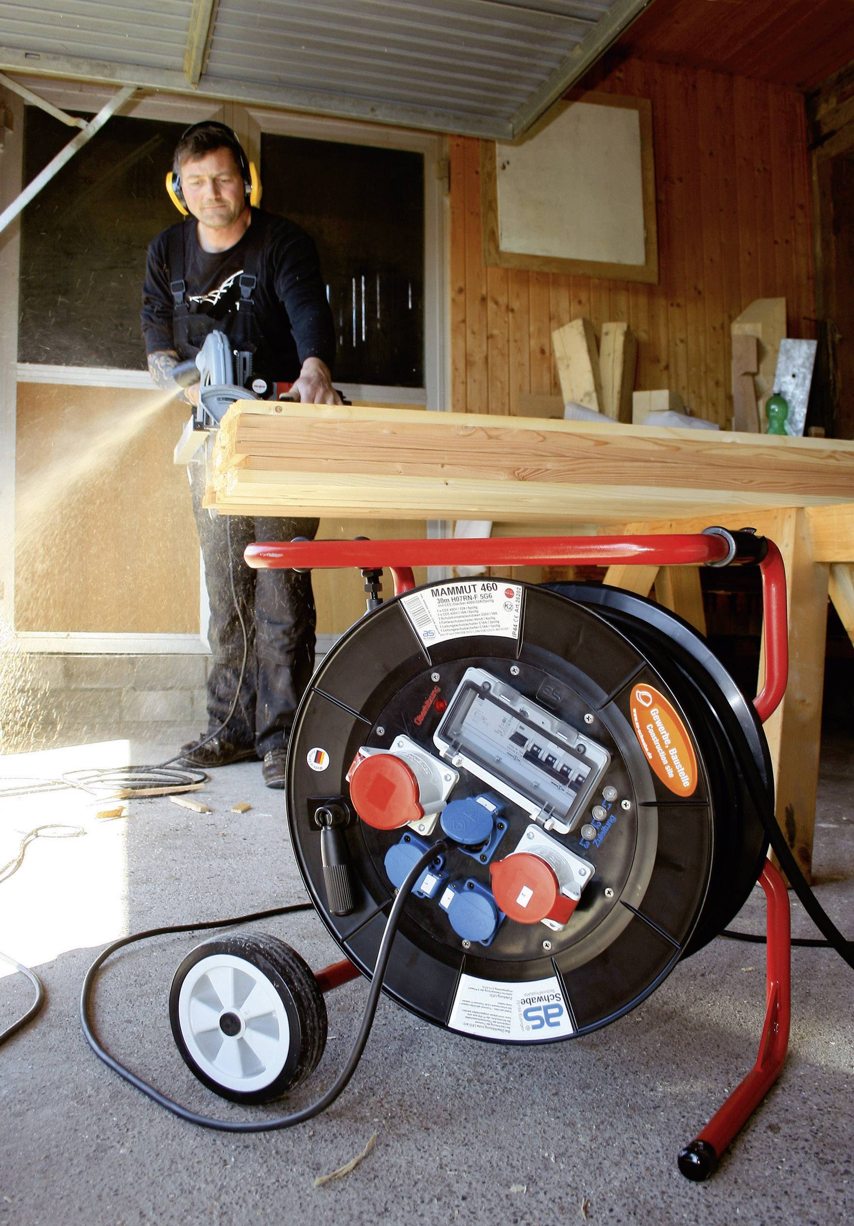 Ein Handwerker schneidet ein Holzbalken mit einer Kreissäge in einer Werkstatt. Im Vordergrund steht ein Kabeltrommel auf einem Rollwagen.