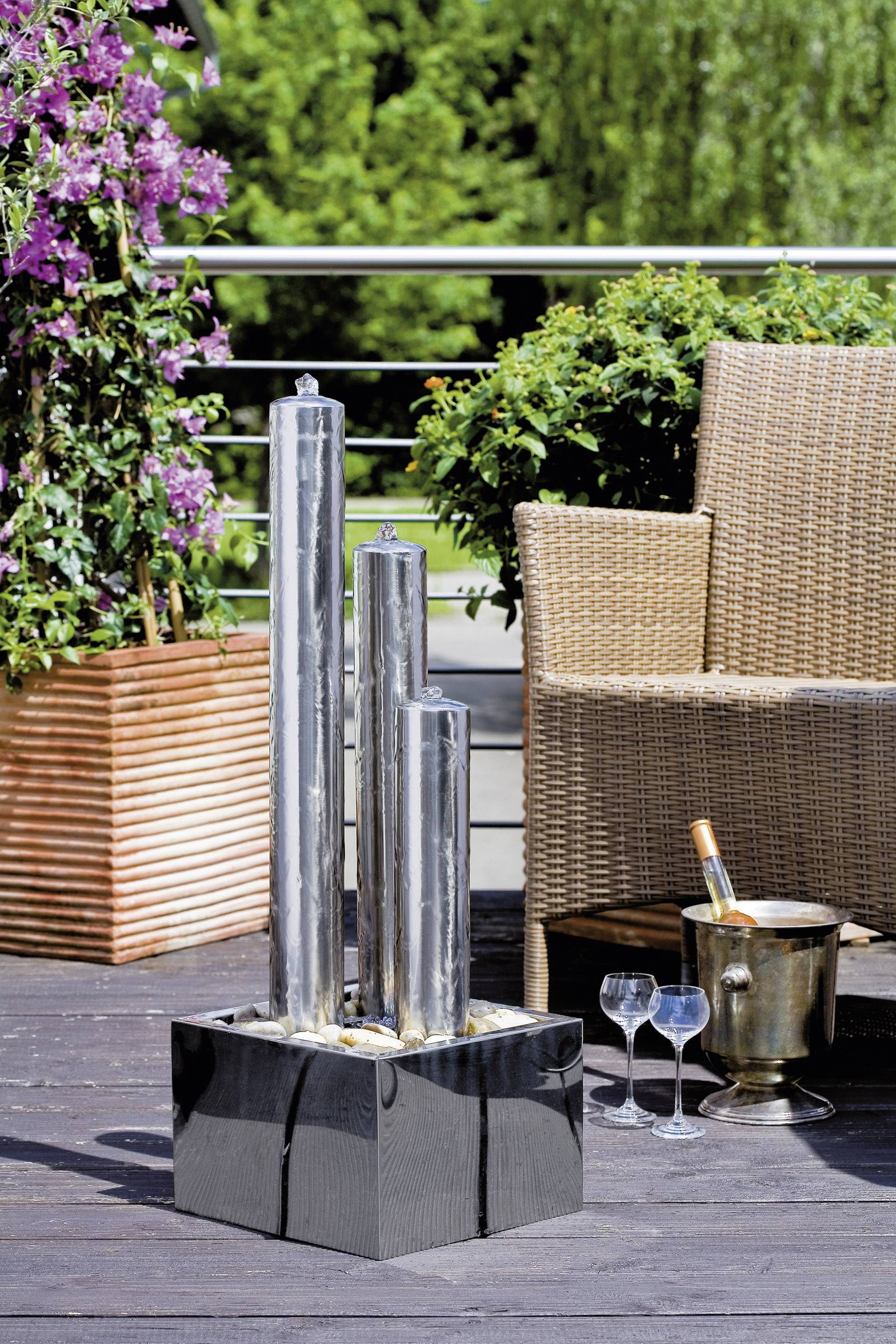 Ein moderner Terrassenbereich mit zwei Stühlen, zwei Gläsern auf dem Boden und einem Eiskübel. Ein hohes Edelstahl-Wasserspiel steht dekorativ im Vordergrund.