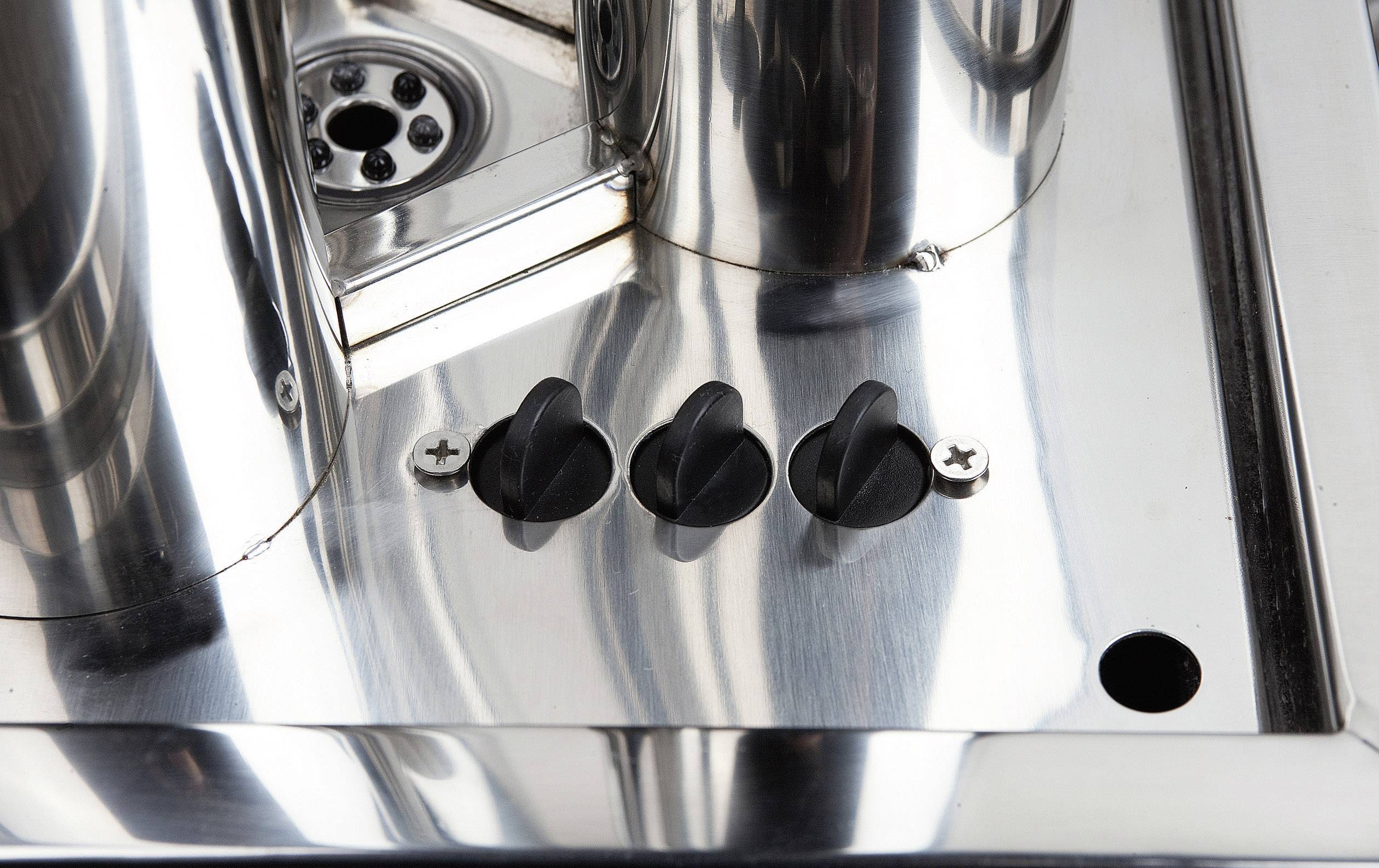 Metallische Oberfläche mit drei schwarzen Drehknöpfen zur Bedienung neben einem runden Abfluss.