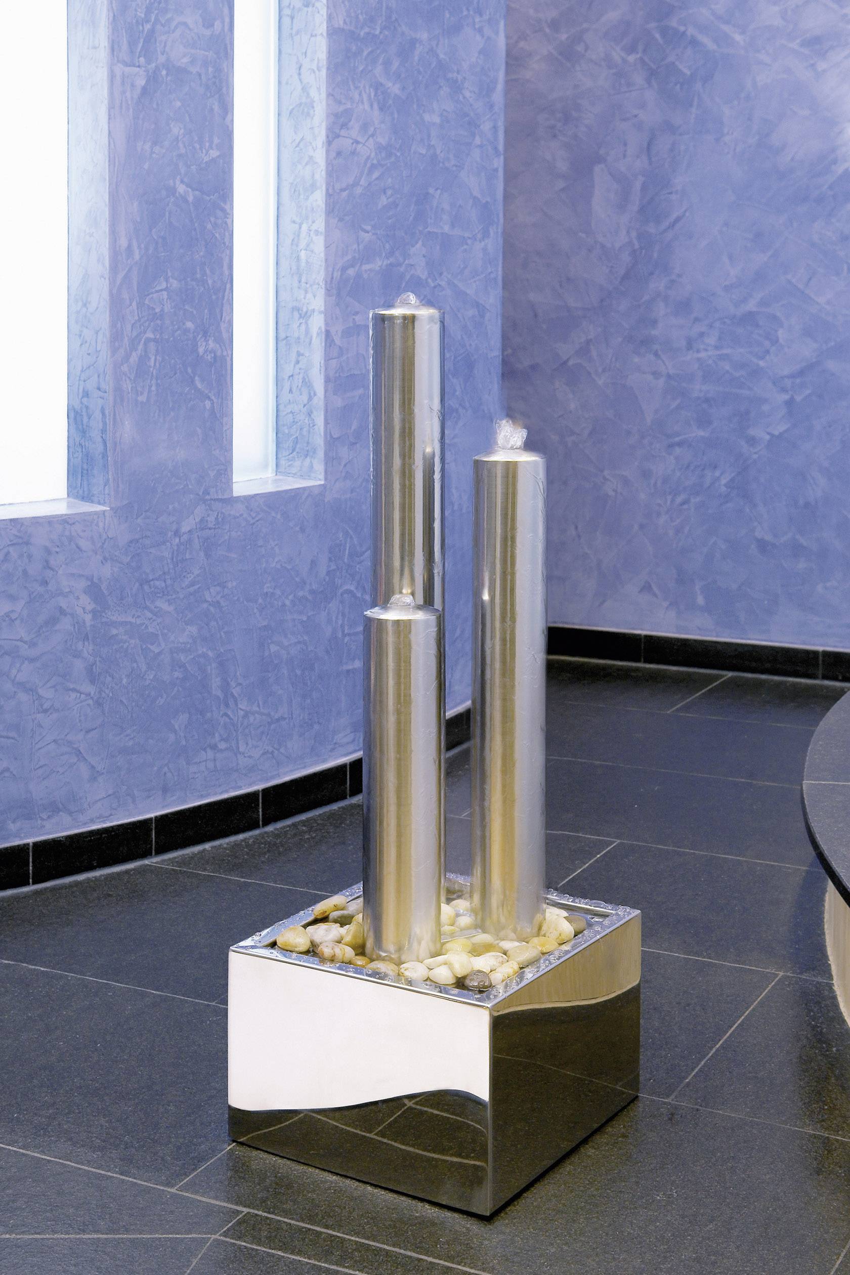 Zwei silberne, zylindrische Wasserfontänen auf einem quadratischen Sockel, umgeben von Steinen, vor einer blau strukturierten Wand.