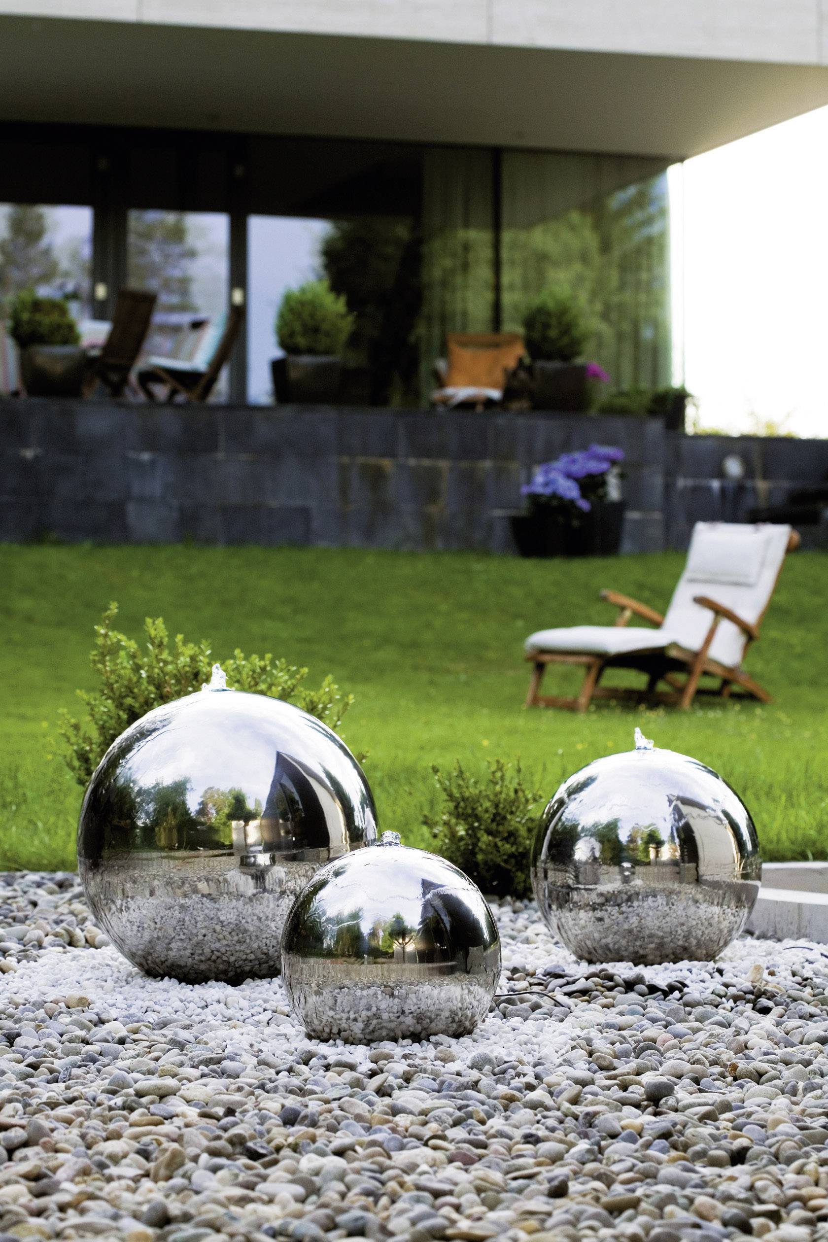 Glänzende Metallkugeln in verschiedenen Größen stehen auf einem Kiesbett vor einem modernen Haus mit grüner Rasenfläche und Liegestuhl.