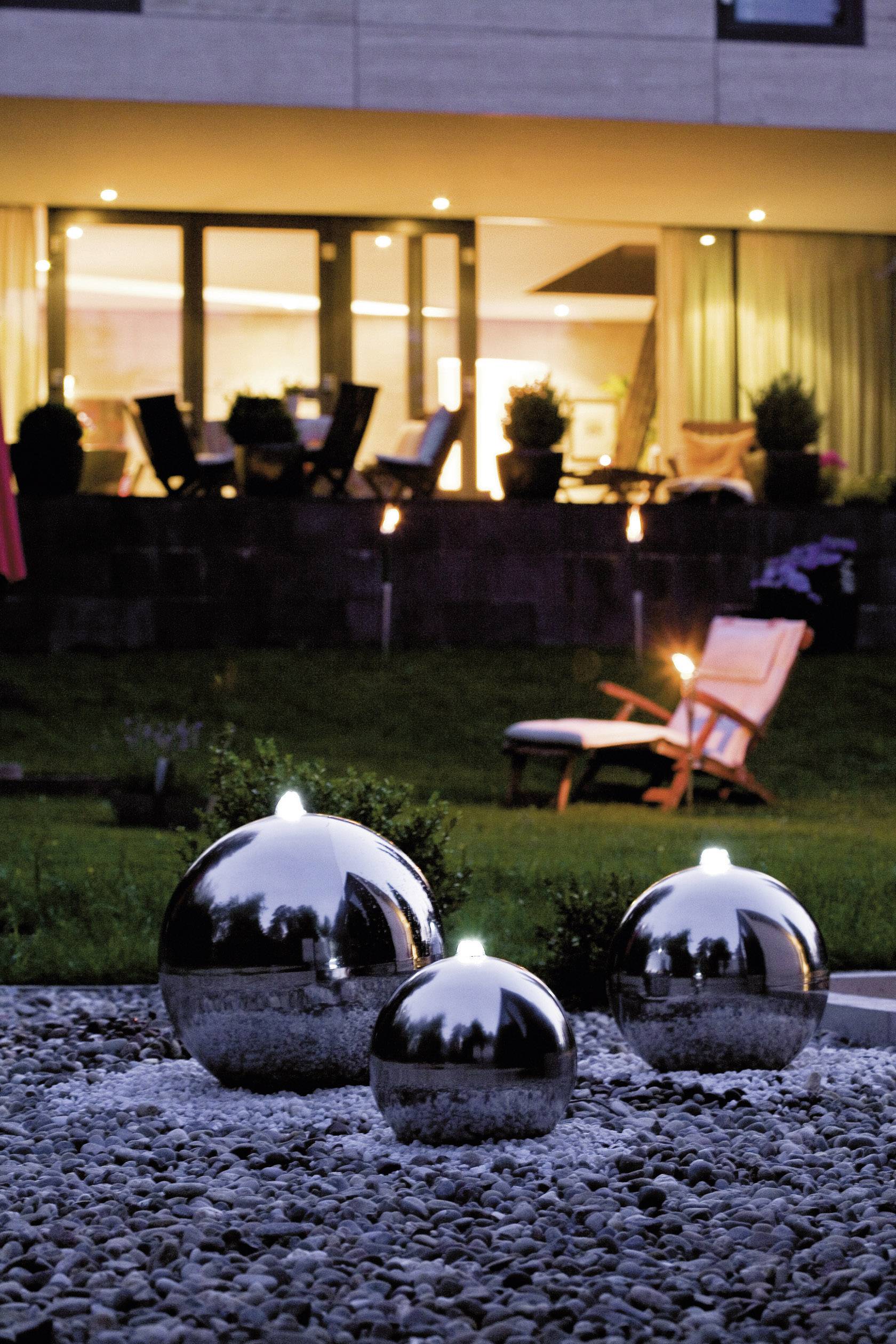 Glänzende Gartenkugeln im Vordergrund, leuchtend im Dunkeln. Im Hintergrund ein beleuchtetes Haus und Gartenmöbel.