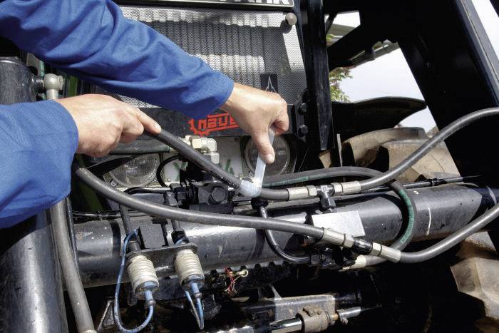 Eine Person repariert oder wartet die Hydraulikschläuche eines Traktors. Die Hände halten Werkzeuge und Schläuche an einer Metallvorrichtung.